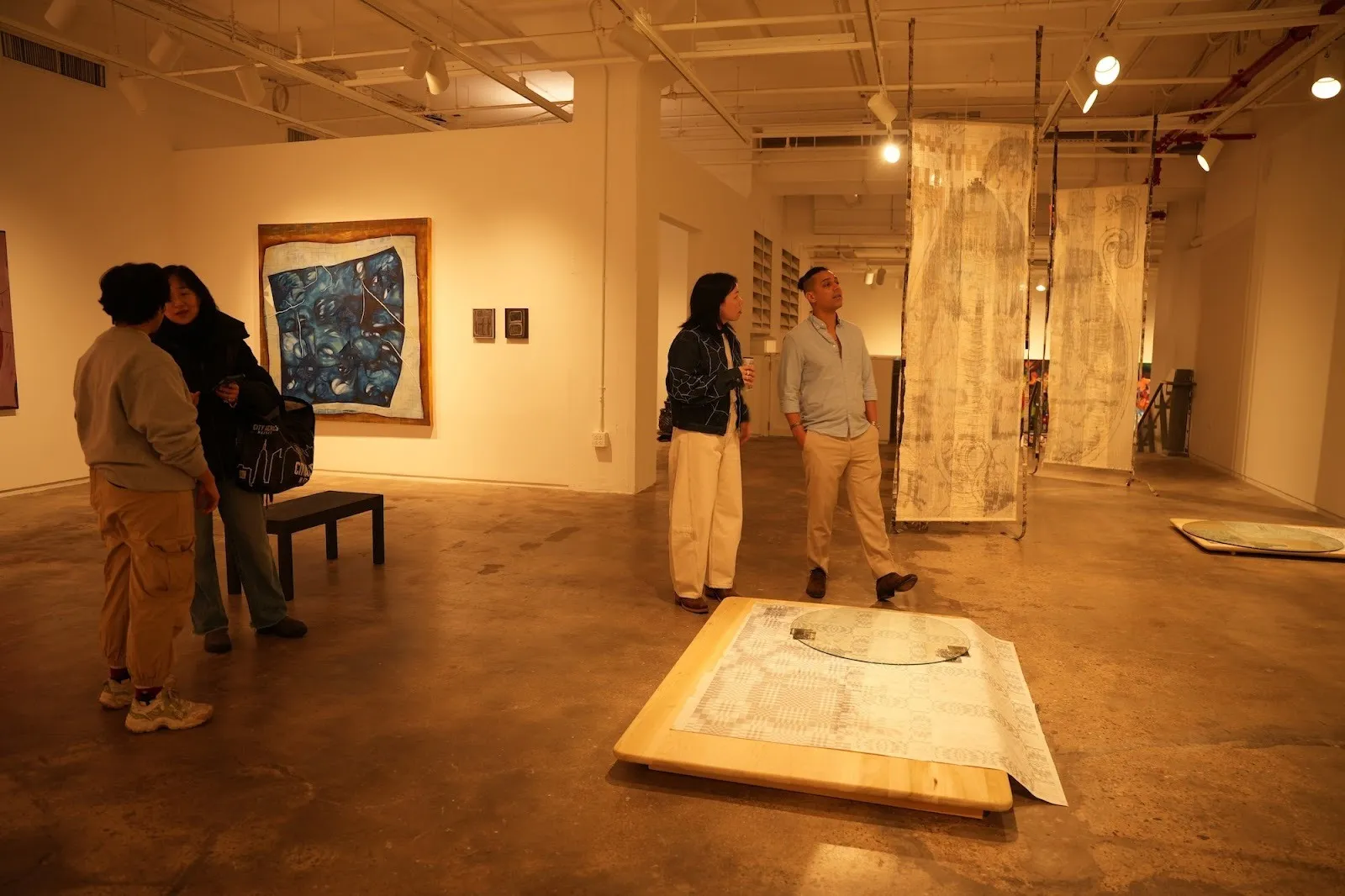 Visitors stand in a contemporary gallery viewing hanging textile works and floor-based sculptures under warm lighting.
