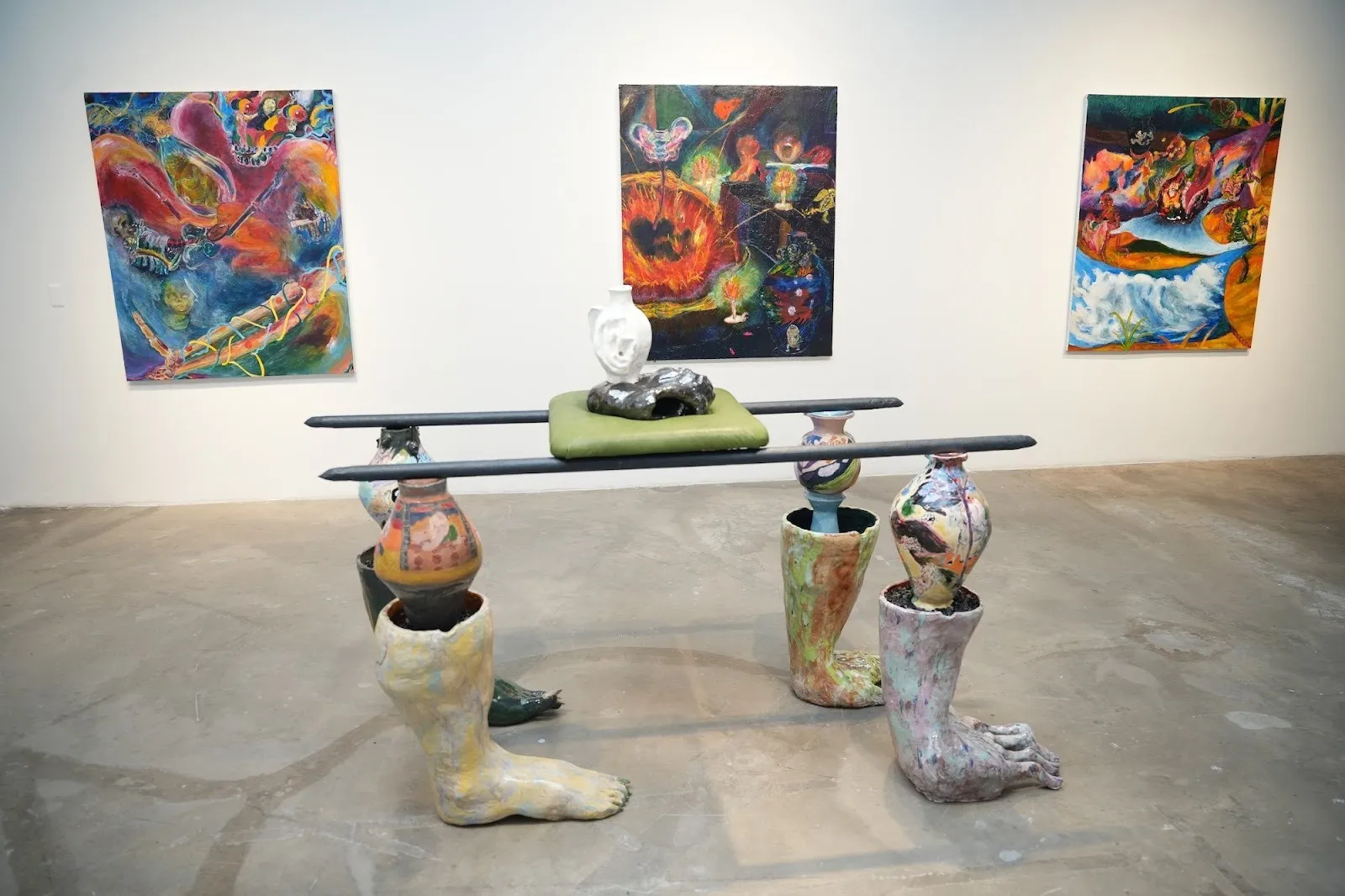 Three colorful, surreal paintings hang behind a sculptural bench made of ceramic legs and vases supporting metal rods.