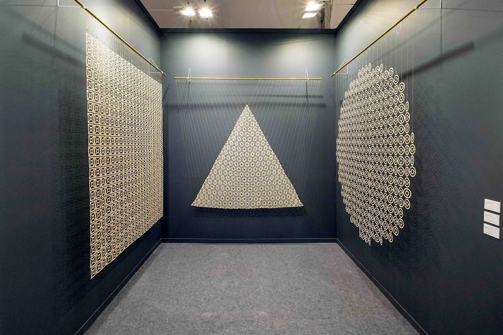 A black-painted booth features tapestries hanging from mounted bars. Each tapestry has a different shape: square, triangle, and circle, and are made from intricate, lace-like weavings.