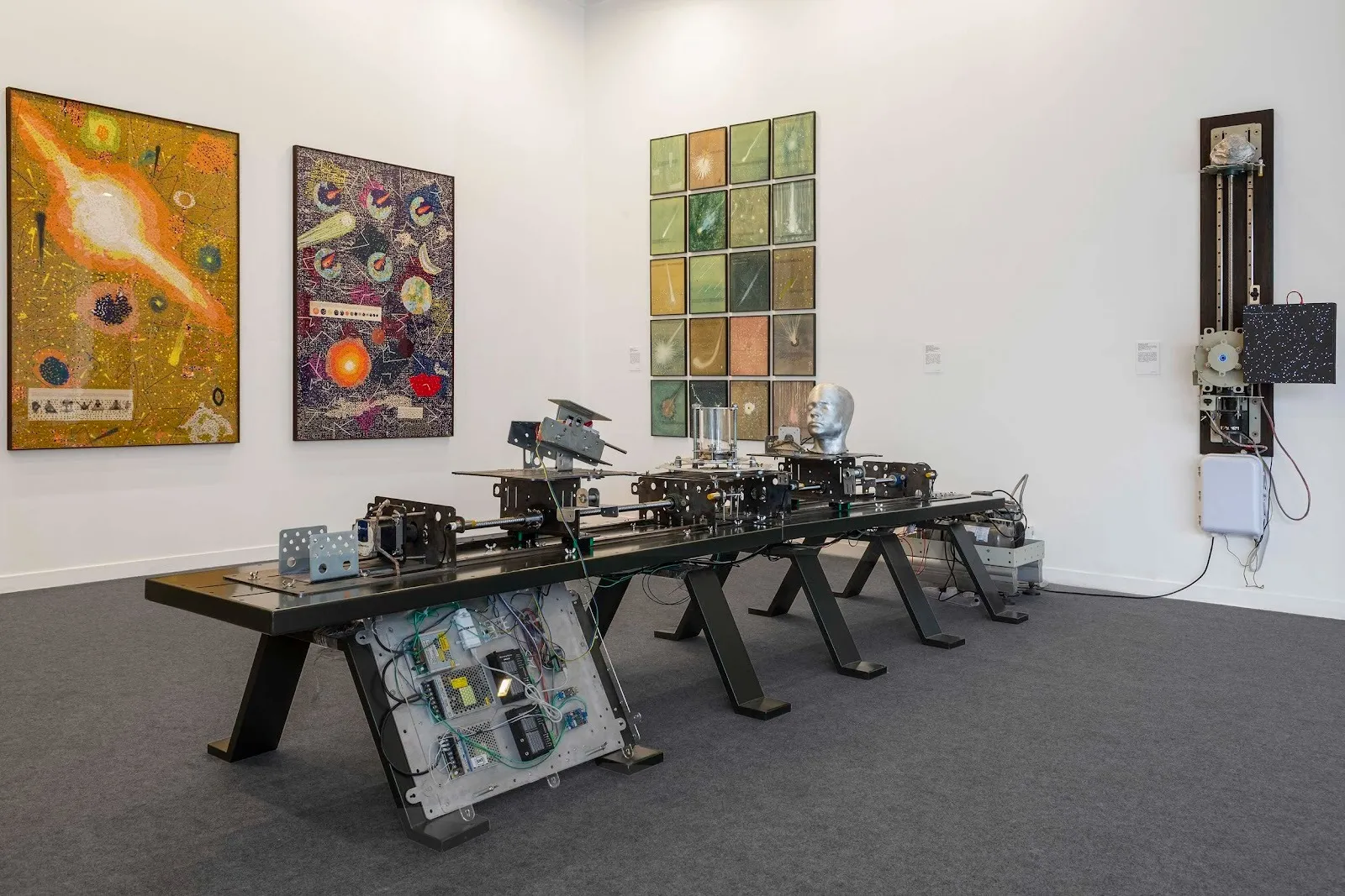 A white booth features multiple hanging 2D works that read like astronomical charts. A large table supports an intricate machine, replete with circuit boards and hydraulics.