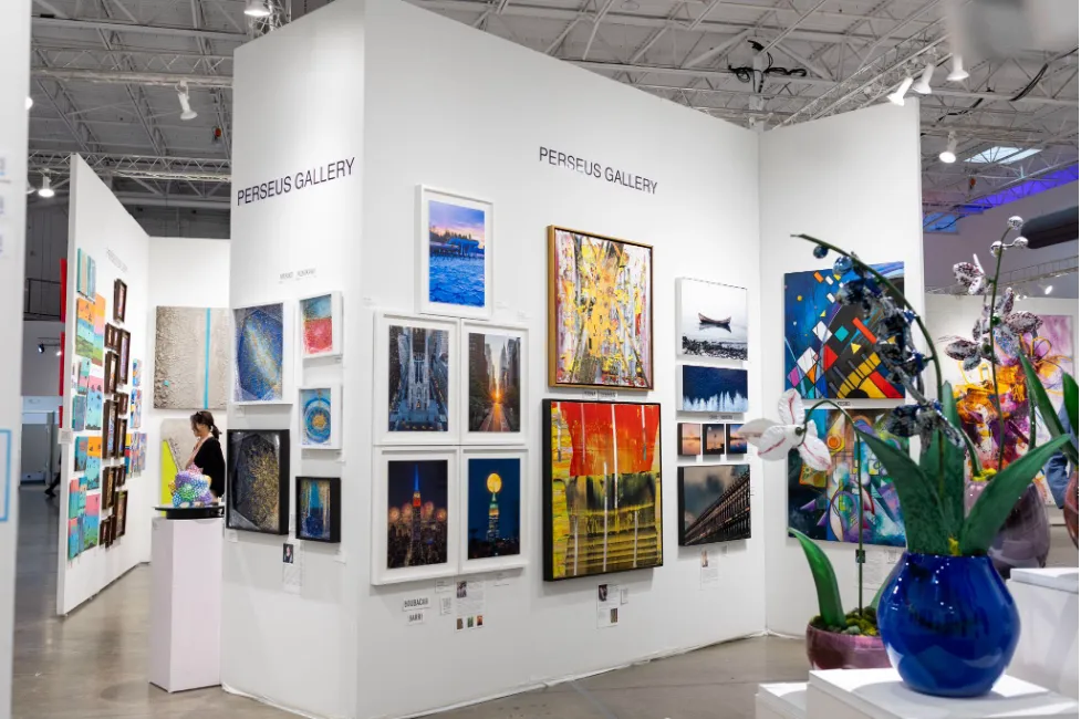 A wide shot of a brightly lit Perseus Gallery art booth, showcasing a large collection of framed photographs, colorful abstract art, and a blue glass vase with flowers in the foreground.