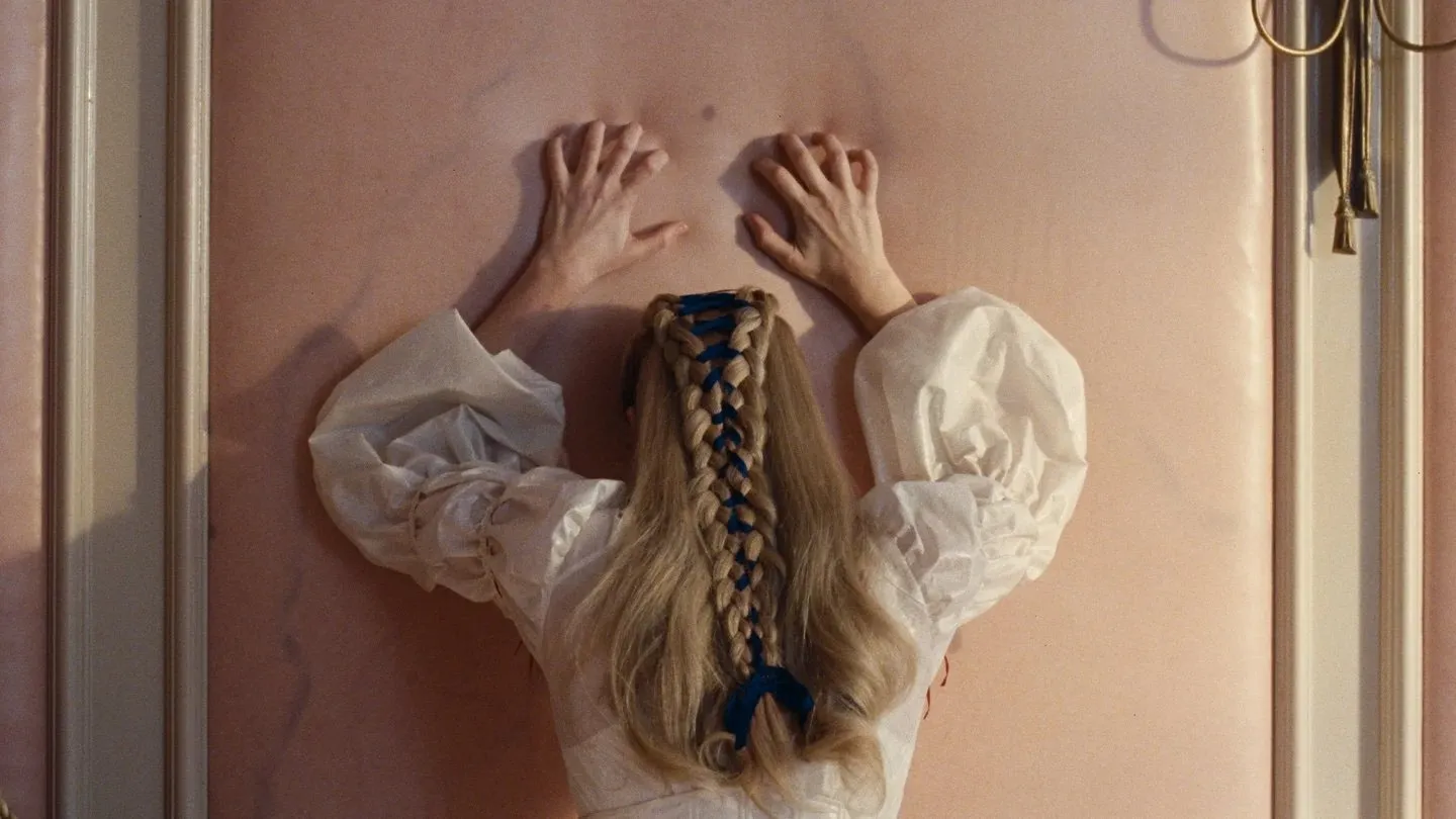 In a flesh-colored room, a woman with elaborately plaited hair in a white dress with puffy sleeves faces a wall and claws at its plush surface.