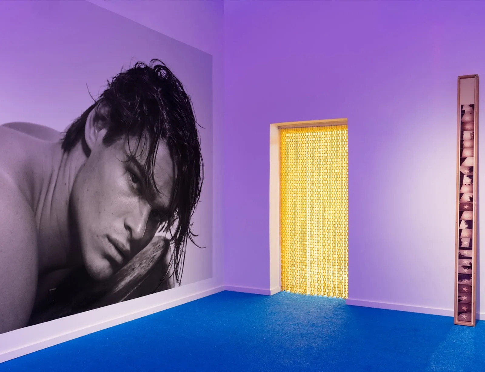 Installation view of a room with blue carpet and purple lighting, featuring a large black-and-white portrait, yellow bead curtains in a doorway, and a narrow vertical artwork.