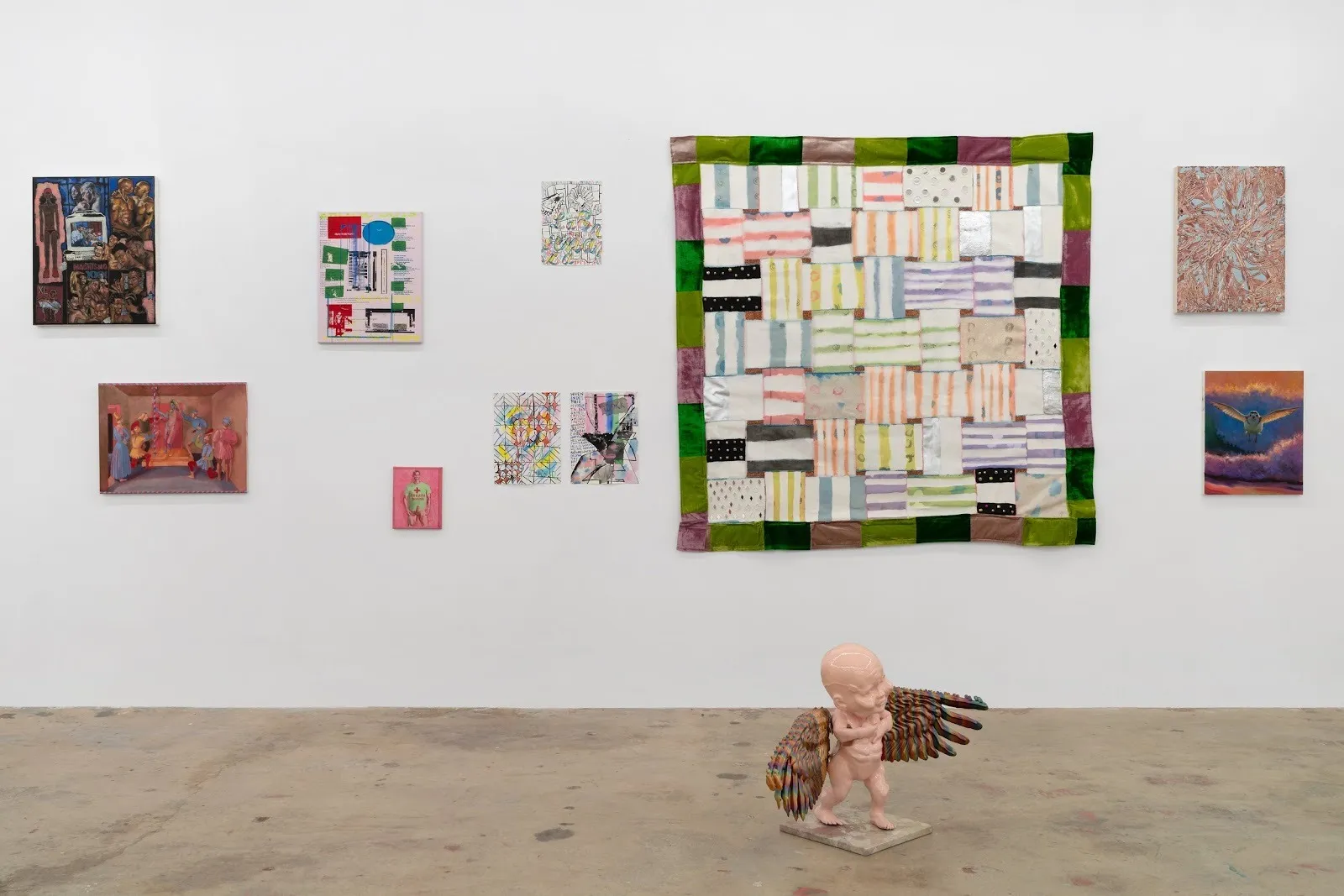 Installation view of a gallery wall with small paintings surrounding a large patchwork textile; a winged baby-like sculpture stands on the floor in front.