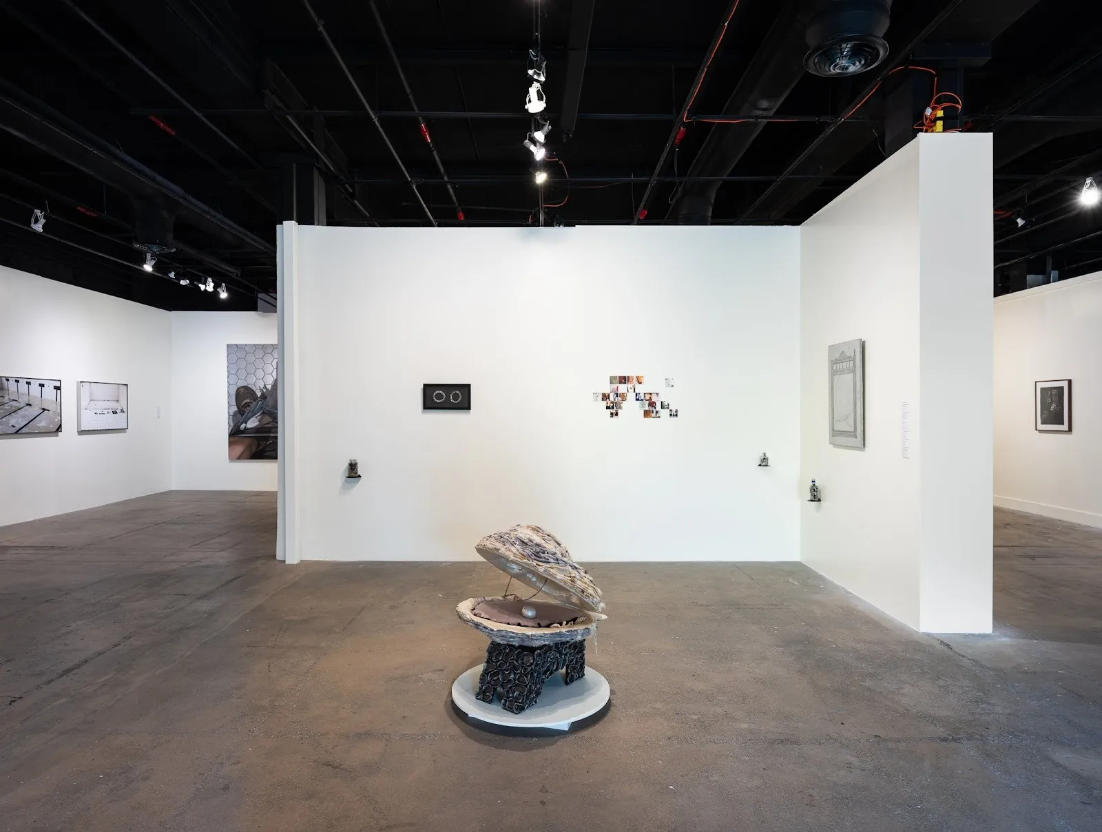 Installation view of a white-walled gallery with framed photographs and small works on the walls; a large clam-like sculpture with a pearl sits on a pedestal in the center.