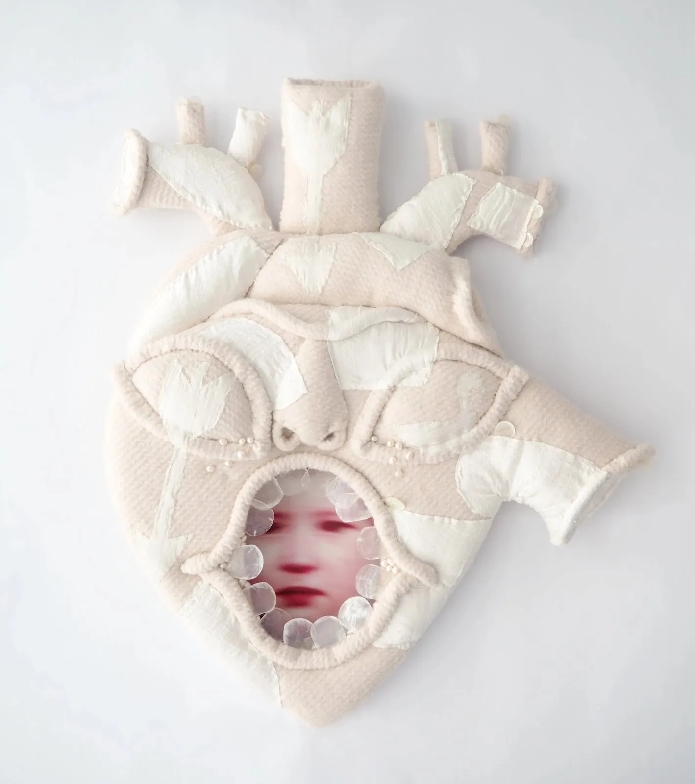 Soft, padded anatomical heart sculpture with stitched fabric panels and a small circular opening revealing a blurred face inside.