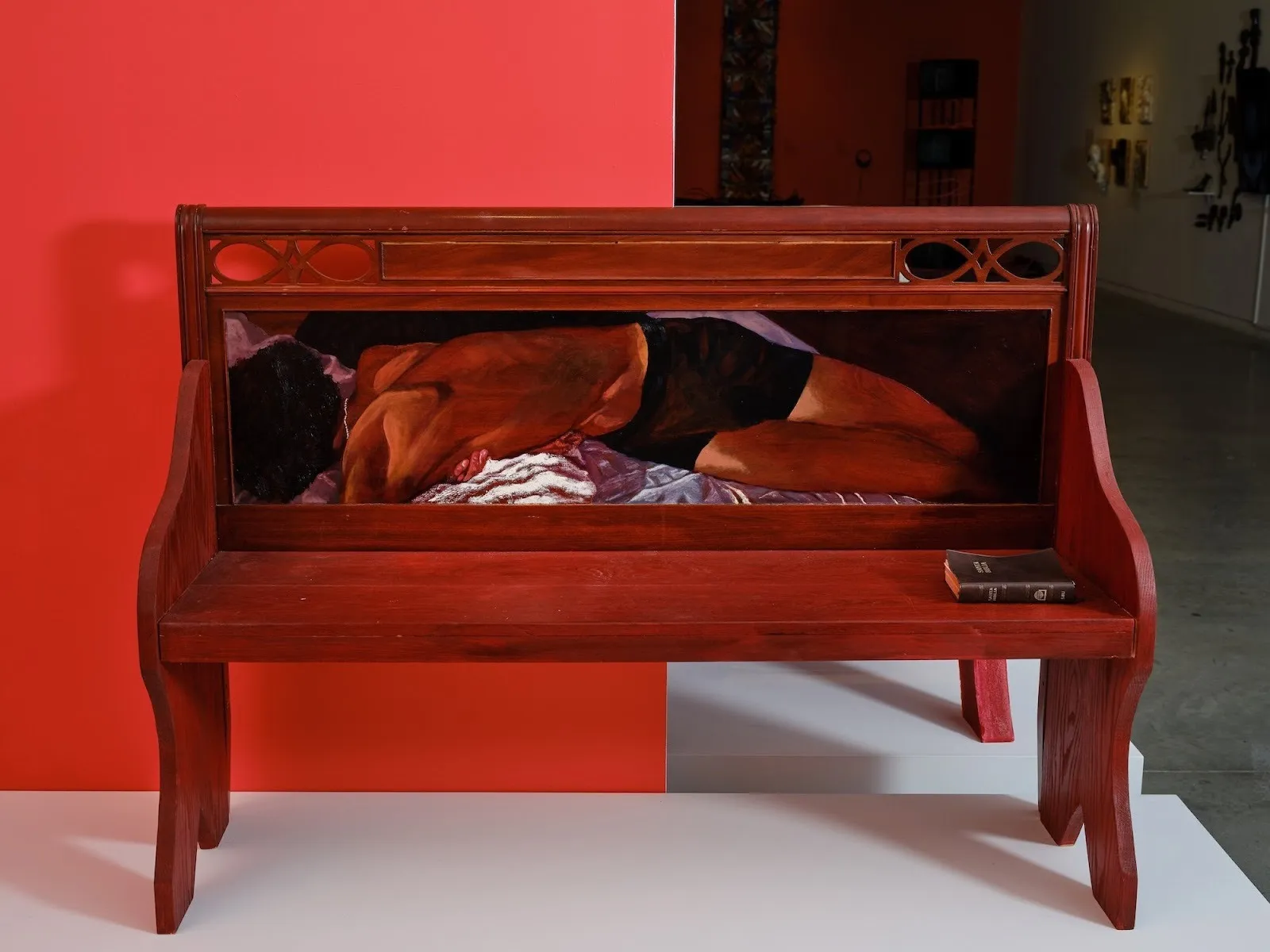 Wooden bench sculpture with a painted figure lying across the backrest panel, set against a red-and-white gallery wall.