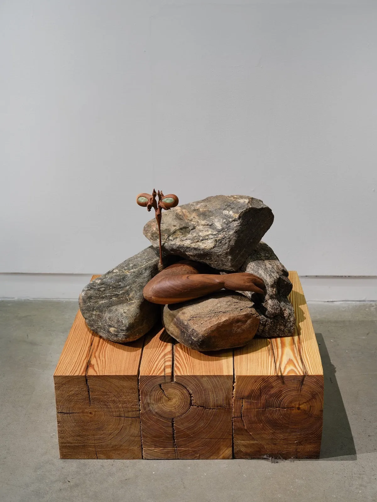 Sculptural arrangement of stacked rocks and carved wood on a wooden block base, featuring a small upright element with eye-like shapes.