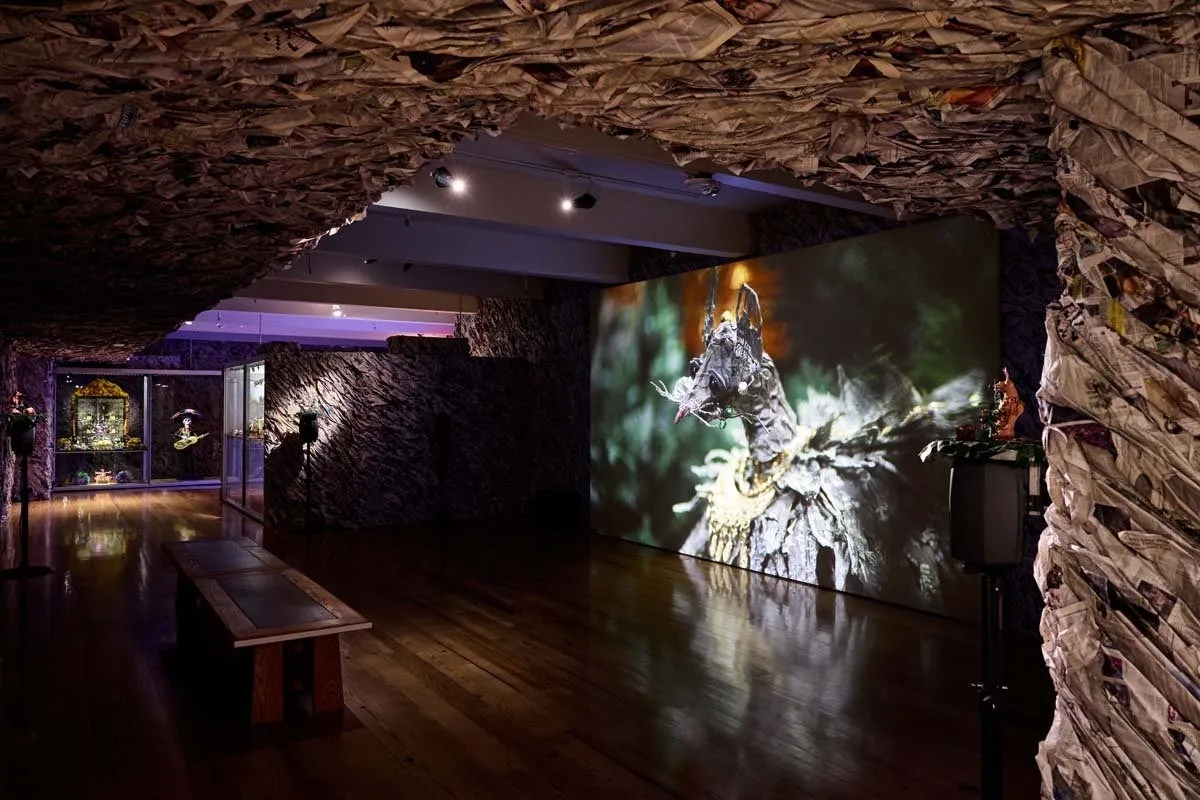 Dark gallery installation with crumpled paper covering walls and ceiling, facing a large projection of a wolf-like figure adorned with gold.