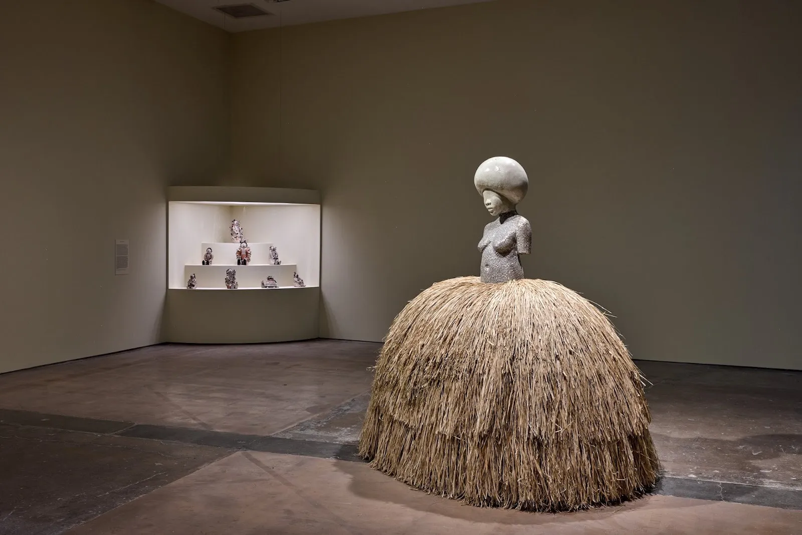 Sculpture of a female torso with a rounded headpiece and a voluminous skirt made of straw, displayed in a dim gallery.