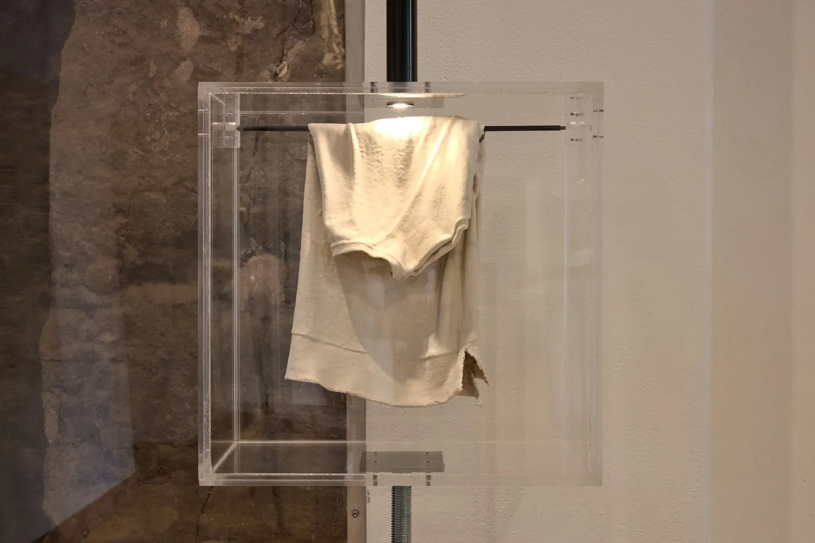 Worn white garment suspended inside a clear acrylic display case mounted on a metal pole.