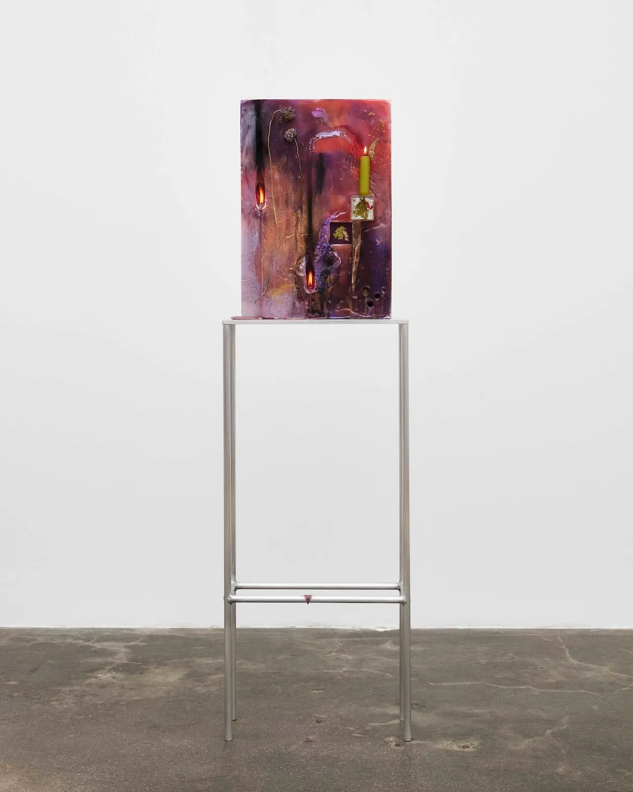 A tall metal table supports a colorful upright vertical surface featuring hues of red and purple. Stuck on the surface are lit candles.