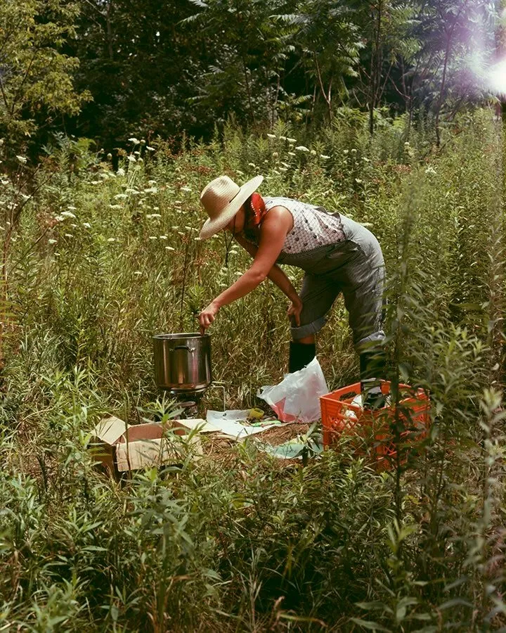 In a densely vegetated field of tall grass and wildflowers, a person in overalls bends over a pot, mixing it. They are surrounded by other miscellaneous supplies, boxes, and crates.