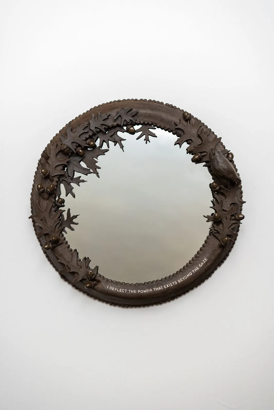 A wall-mounted circular mirror is framed by a thick, brown frame decorated with sculpted leaves, acorns, and a bird from the same frame material. On the bottom, text curved with the mirror reads "I REFLECT THE POWER THAT EXISTS BEYOND THE GAZE."