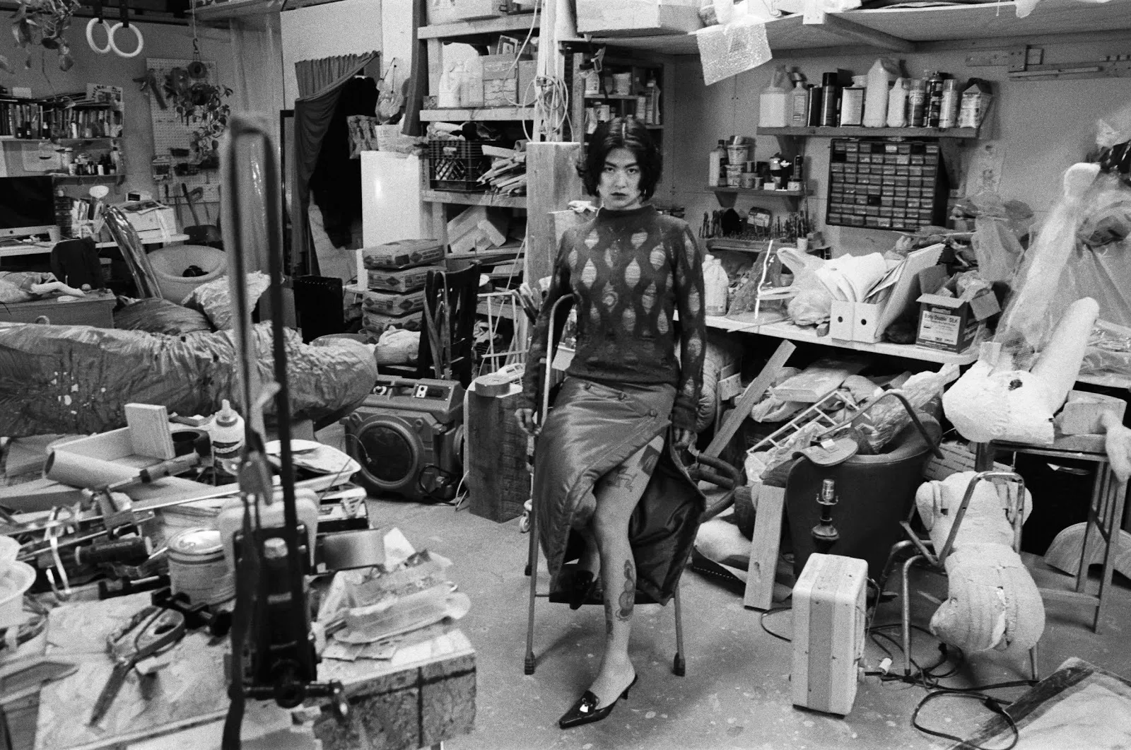 A black and white shot of a person leaning on a stepladder in a densely cluttered studio. The person stares directly at the camera and is surrounded by tools, mannequins, furniture, and other miscellaneous creative objects.