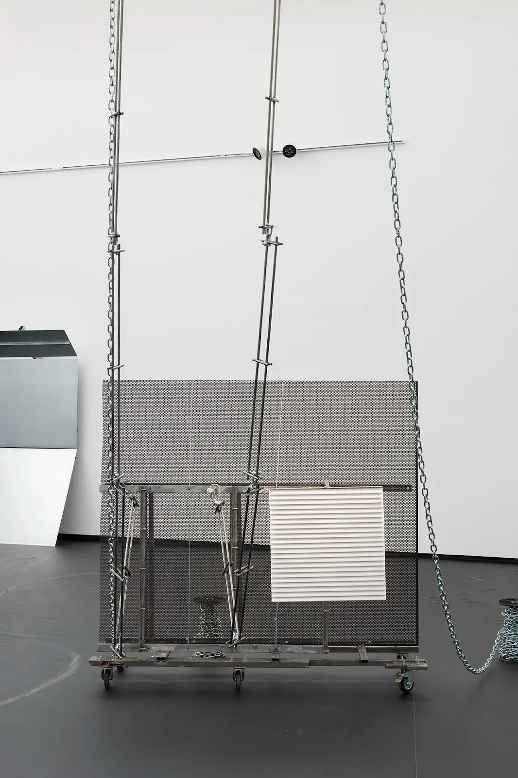 Metal sculpture on wheels with mesh panel, chains, rods, and a white corrugated panel suspended in a gallery space.