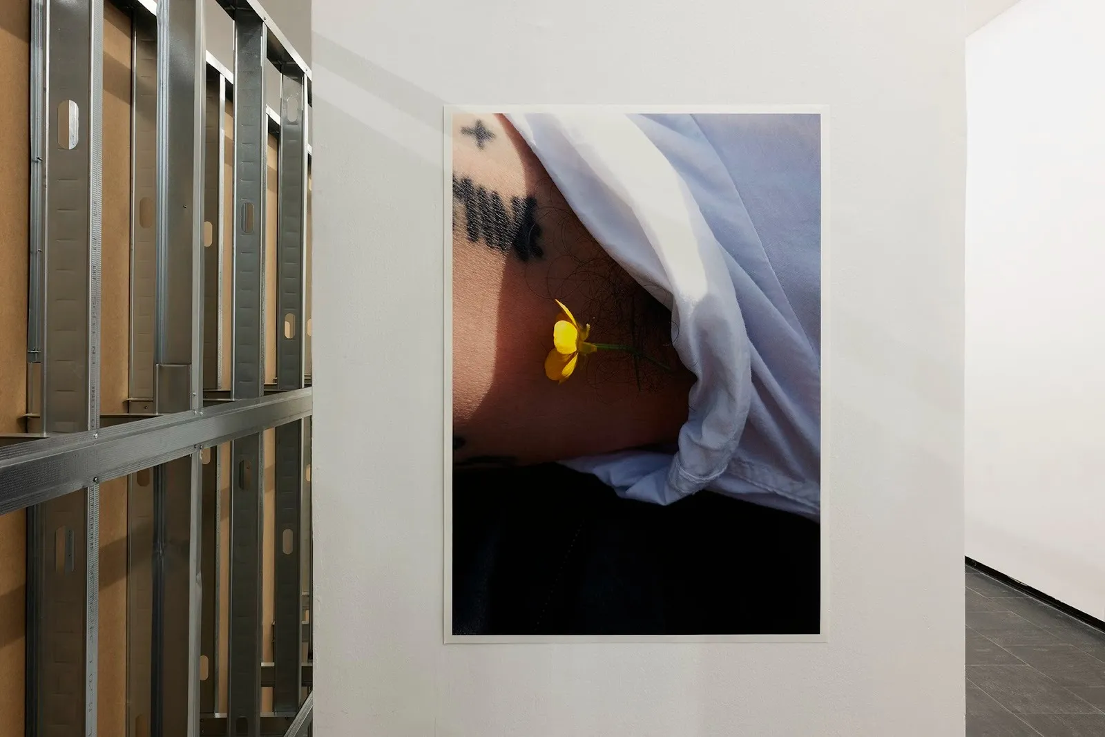 Photograph of a person’s torso partially covered by white fabric, with a small yellow flower resting against the skin near a tattoo.