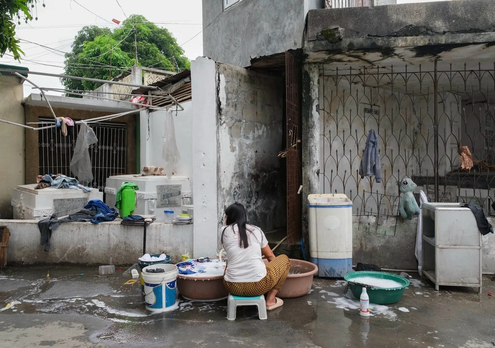 Woman washing clothes in plastic basins beside weathered cemetery tombs and household items in a small courtyard.
