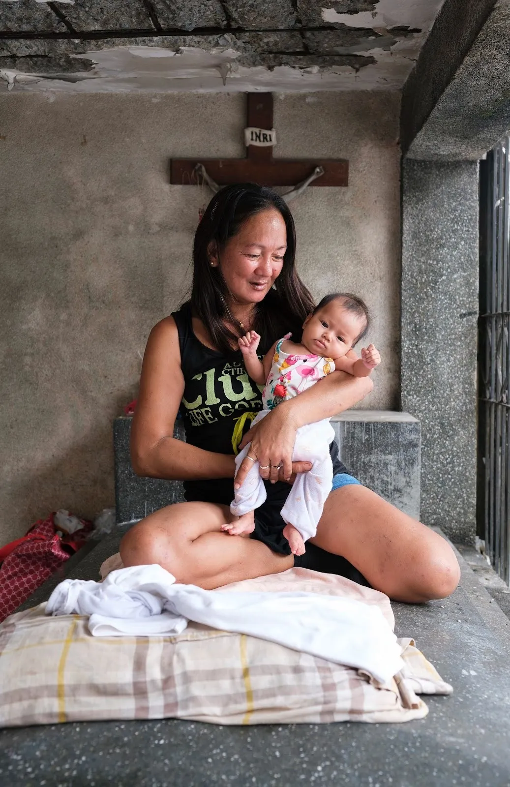 Woman sitting inside a concrete tomb holding a baby, with a crucifix mounted on the wall behind them.