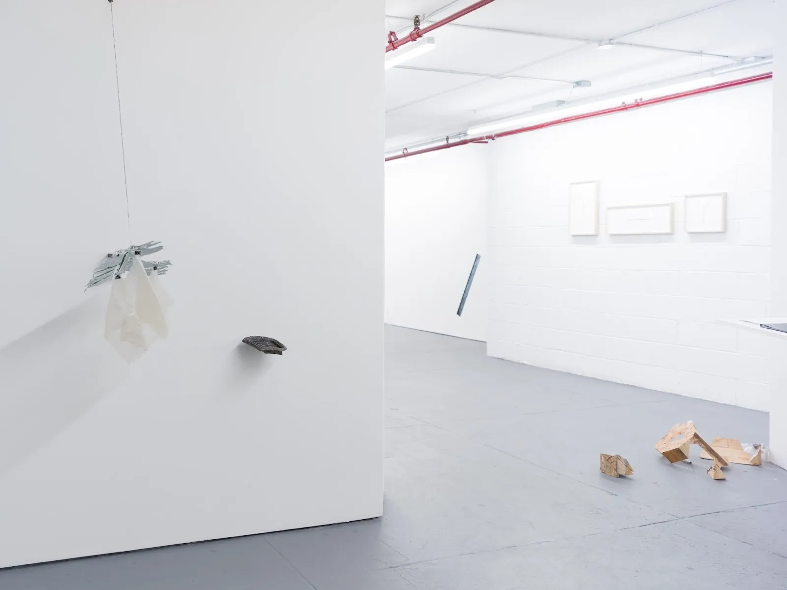 Gallery installation view with hanging translucent sculpture, wall-mounted piece, and scattered wooden fragments on the floor.