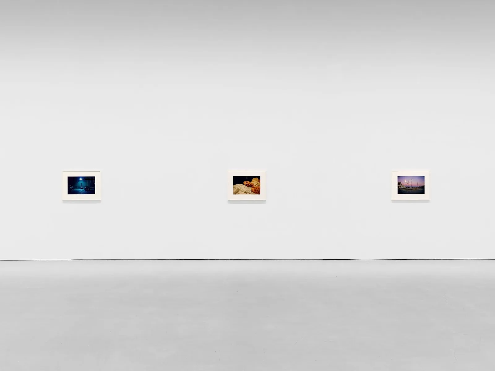 Gallery wall with three framed photographs depicting a nighttime street scene, a person sleeping, and a lit roadside at dusk.