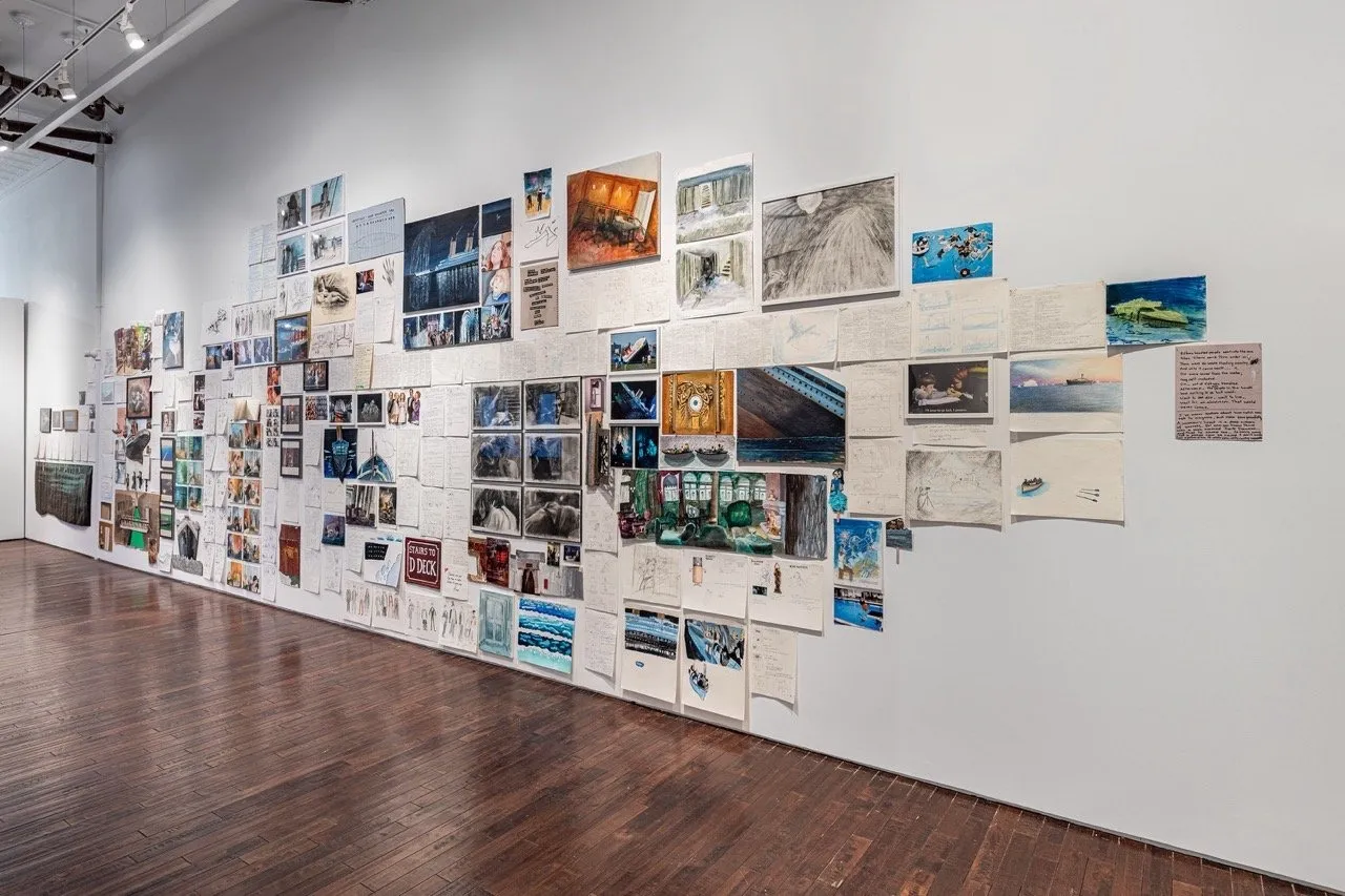 Gallery installation view of a floor-to-ceiling collage of drawings, photographs, sketches, and handwritten notes covering an entire white wall.
