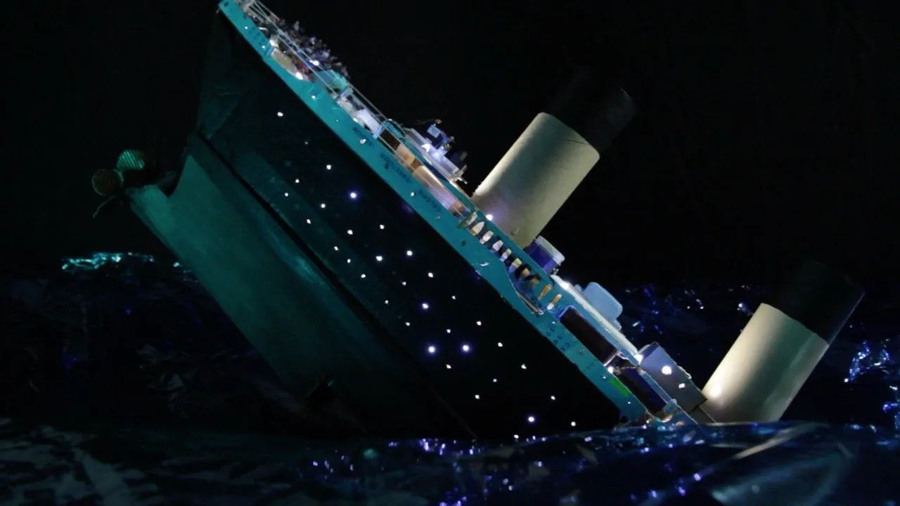 A illuminated model of the Titanic tilting steeply into dark, glittering water against a black background.