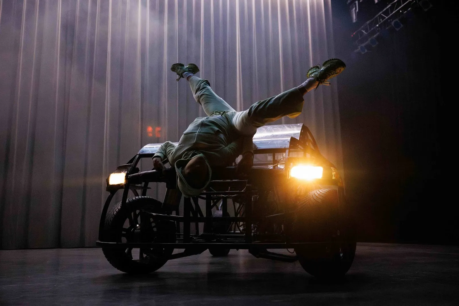 A performer in a full cloth mask and a grey sweatsuit dances over the frame of a car with headlights. The dancer hoists themselves over the hood of the car on their back, positioned upside down with their legs in the air.