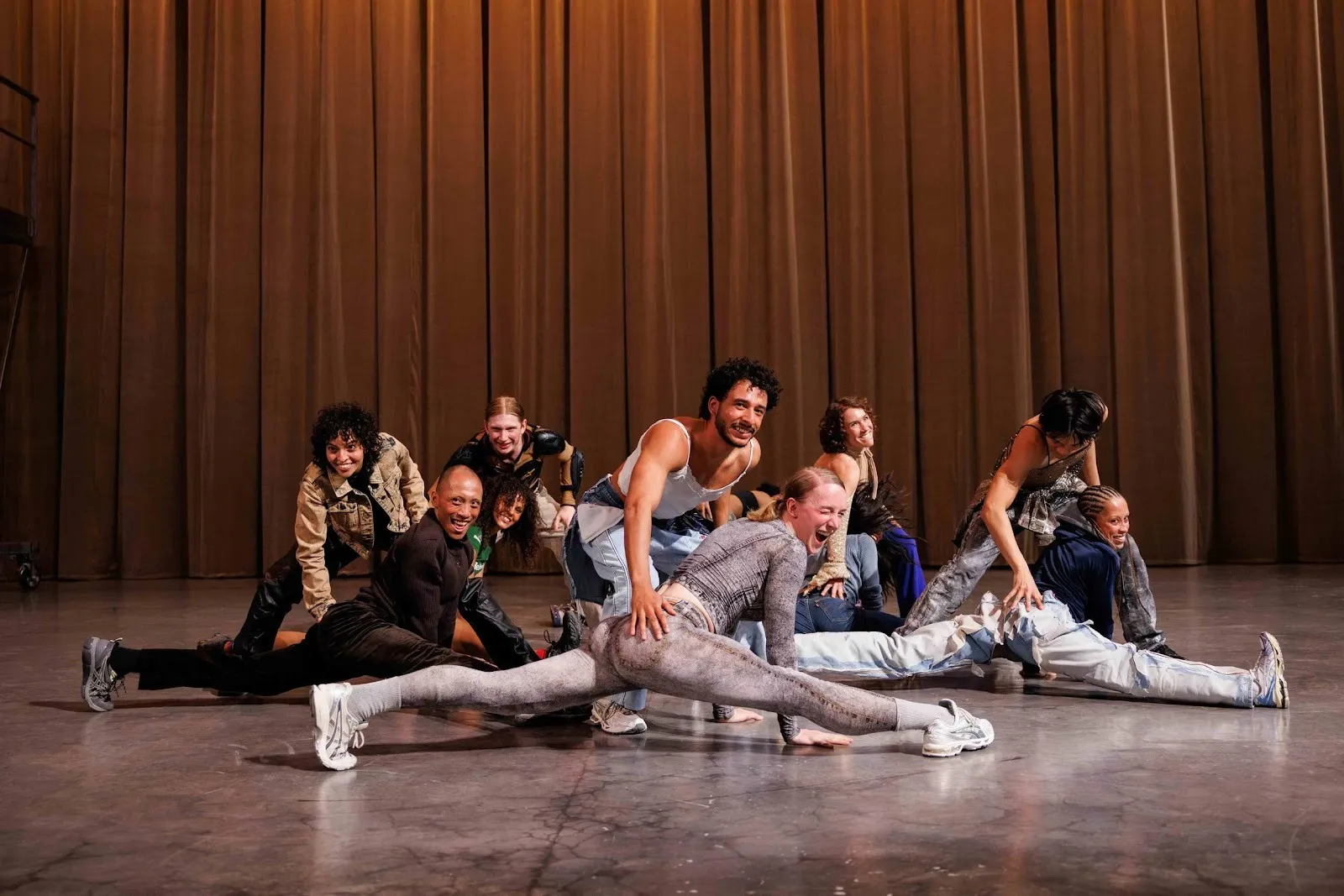 On a minimal stage with a gold back curtain, a group of performers smile as they replicate an exercise class-like scenario, with pairs of dancers mimicking instructors helping their student with a split exercise.