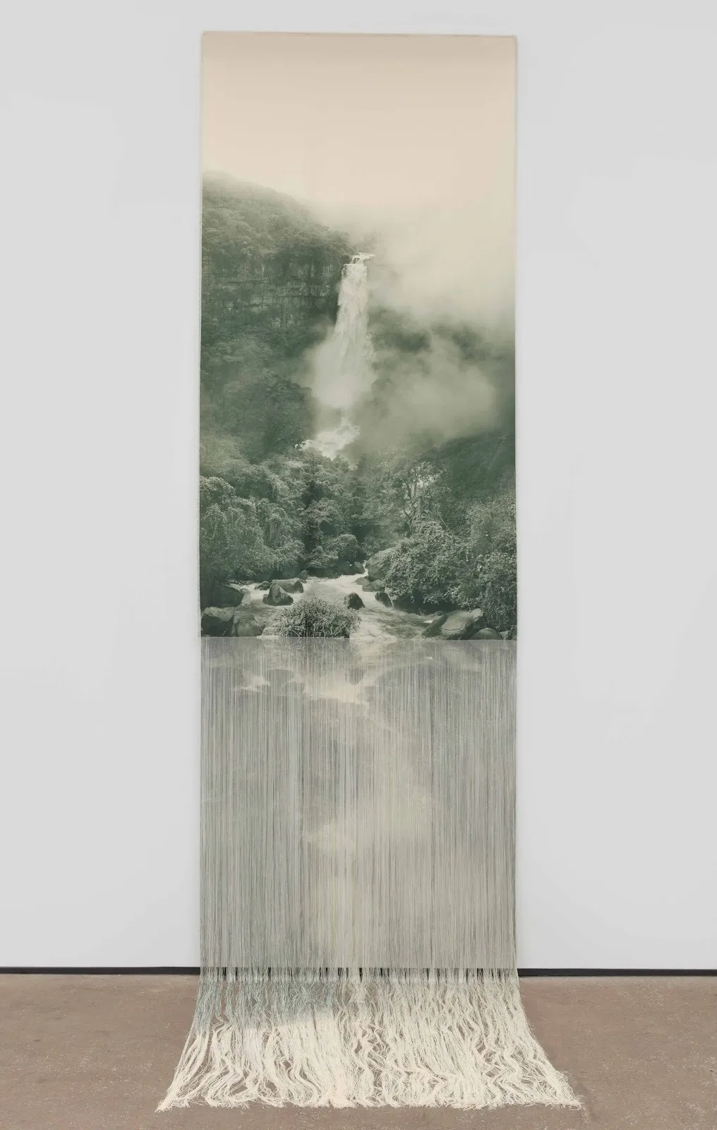 A long, vertical wall work is made of fabric printed with dye. The bottom half of the work is unraveled into threads that spill onto the floor. The printed image depicts a photograph of a green and beige mountainous jungle landscape and waterfall.