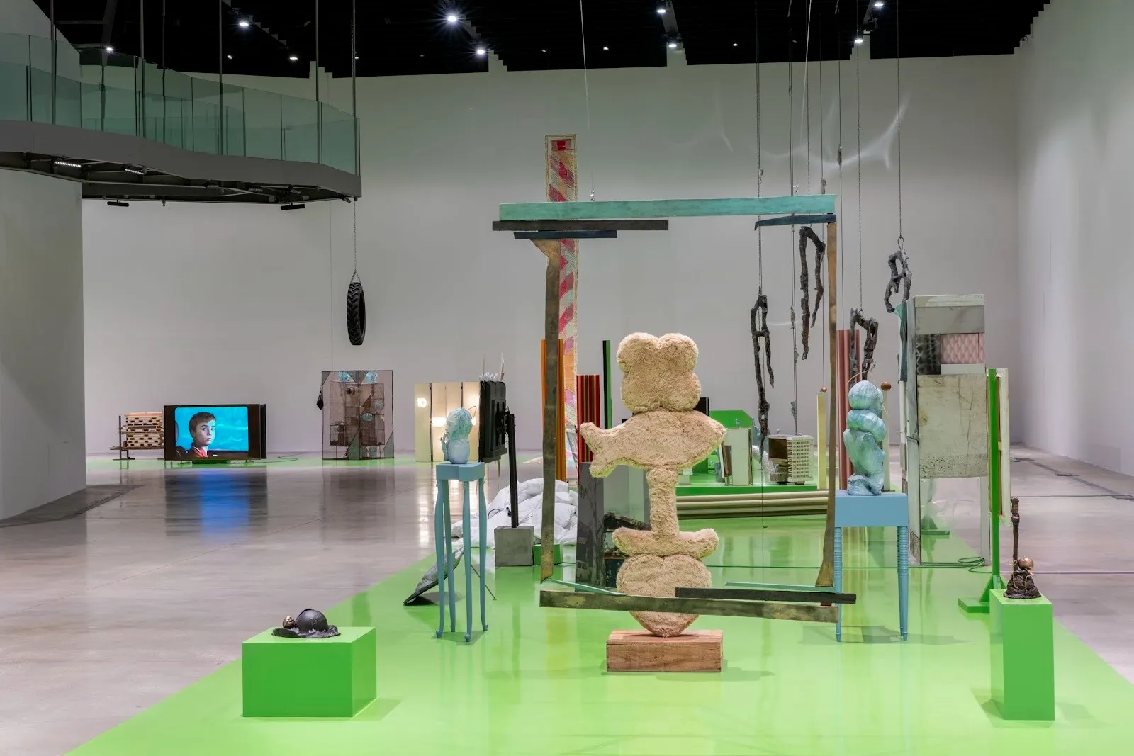 A large, white gallery room with a second-story glass balcony has a strip of green flooring running centrally throughout the room. On the green strip is a collection of sculptures, all visually distinct, but grouped closely together.