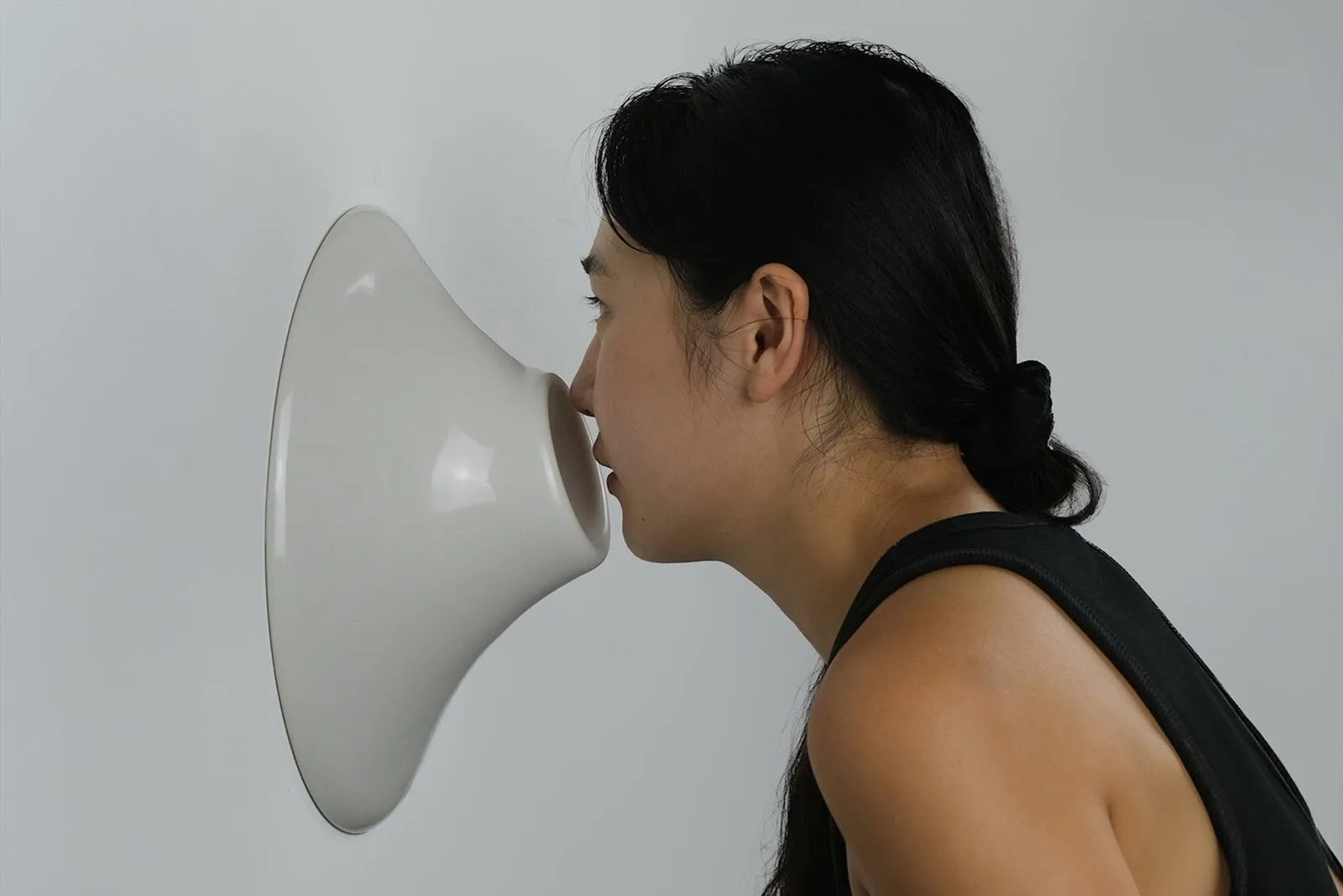 A woman in profile presses her face into a smooth, white, wall-mounted sculptural form against a plain background.