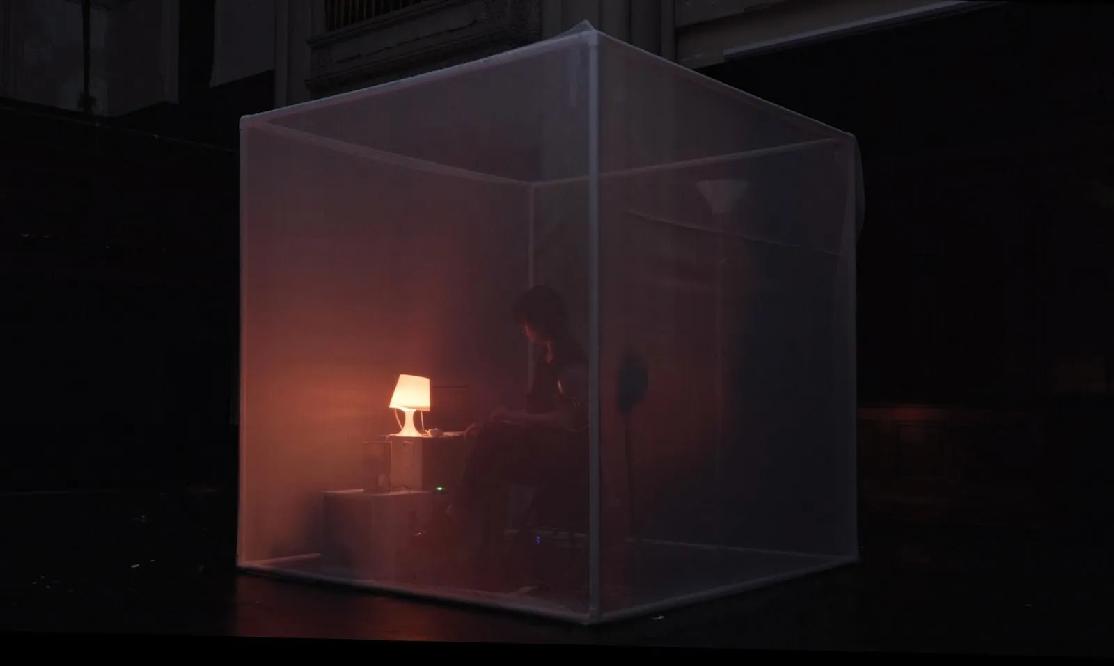 A person sits inside a semi-transparent cube lit by a warm table lamp, creating an intimate, dimly lit performance environment.