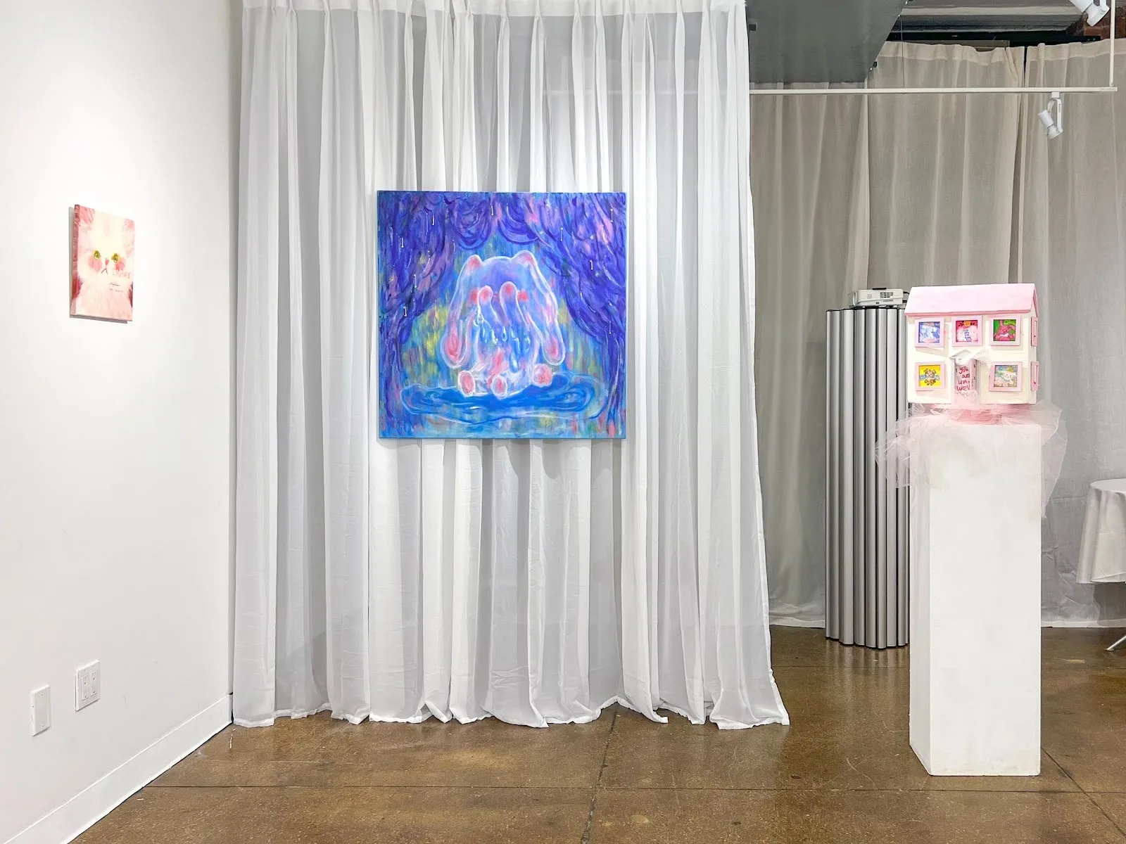 Exhibition space with a sheer white curtain backdrop, a blue abstract painting centered, and a small pink house sculpture displayed on a pedestal to the right.