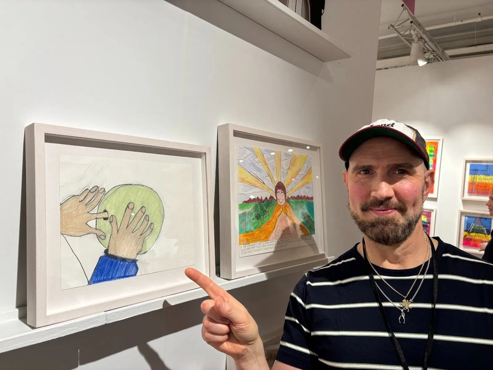 Man in a cap points at framed drawings on a gallery wall, including an image of hands holding a green sphere.