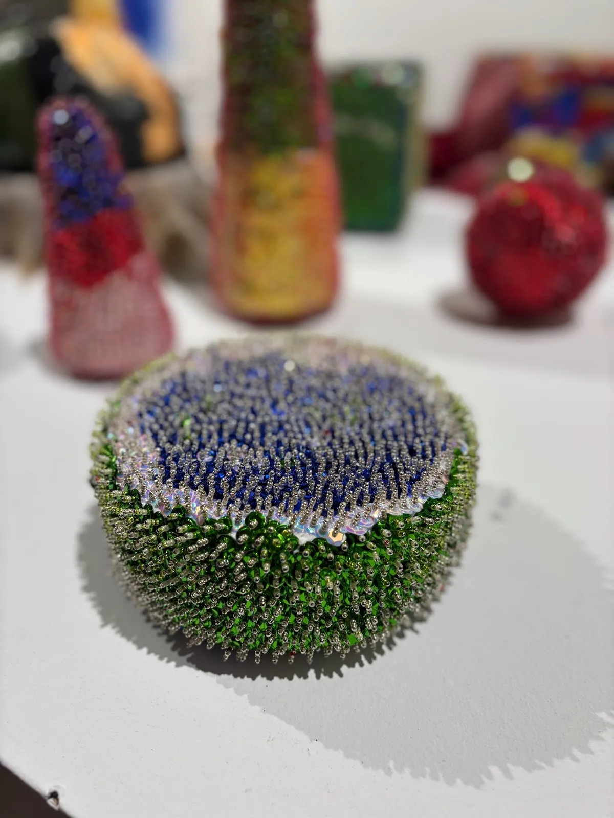 Close-up of a round, spiky beaded sculpture with green sides and a blue center, surrounded by other blurred beadwork.