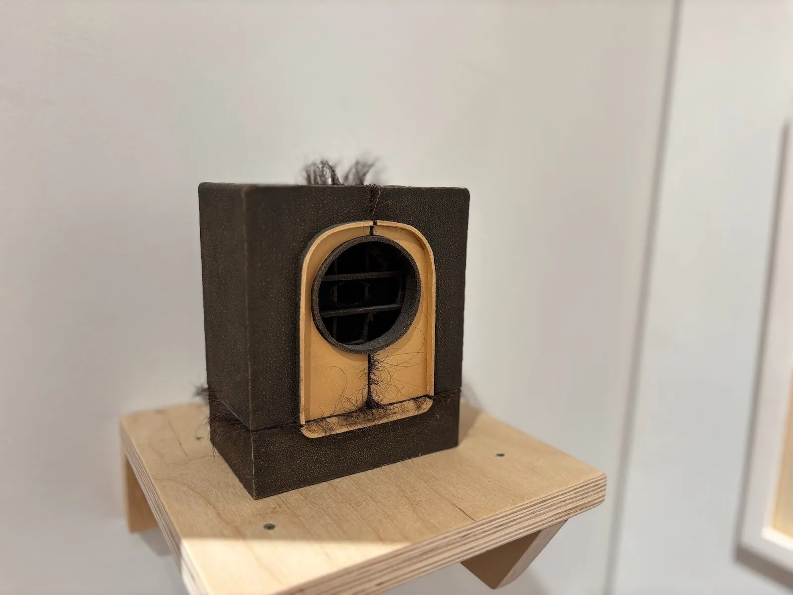 Small sculptural object resembling a vintage radio, wrapped in dark material with strands of hair emerging from its seams.