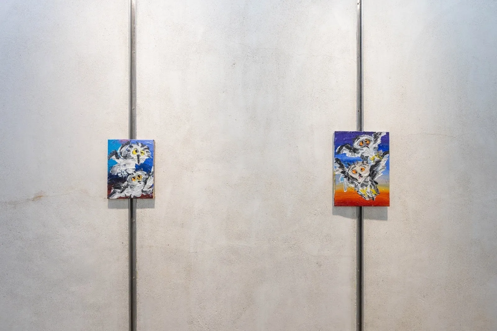 Two small, expressive owl paintings mounted on a concrete wall, each featuring loose brushwork and bright, contrasting backgrounds.