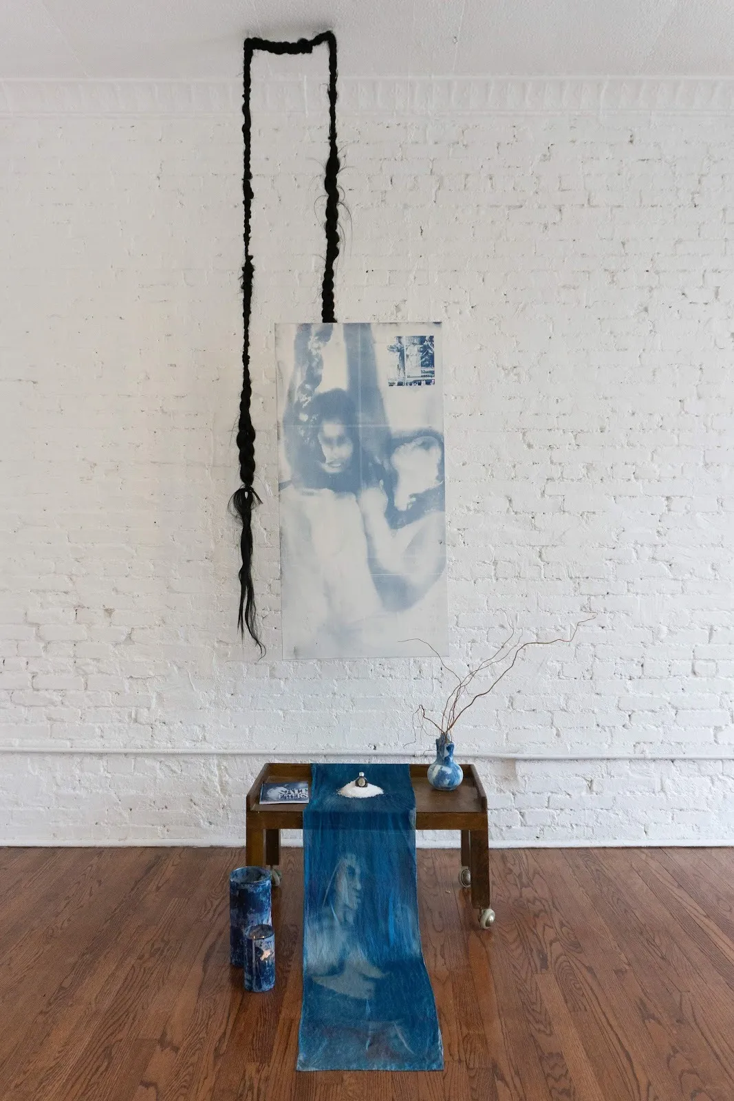 In a white brick-walled room with hardwood floors, an installation is comprised of distinct parts: a long rope of braided hair, an image printed in blue on silk, a small wheeled table on the floor with more printed fabric, a blue vase, book, and more
