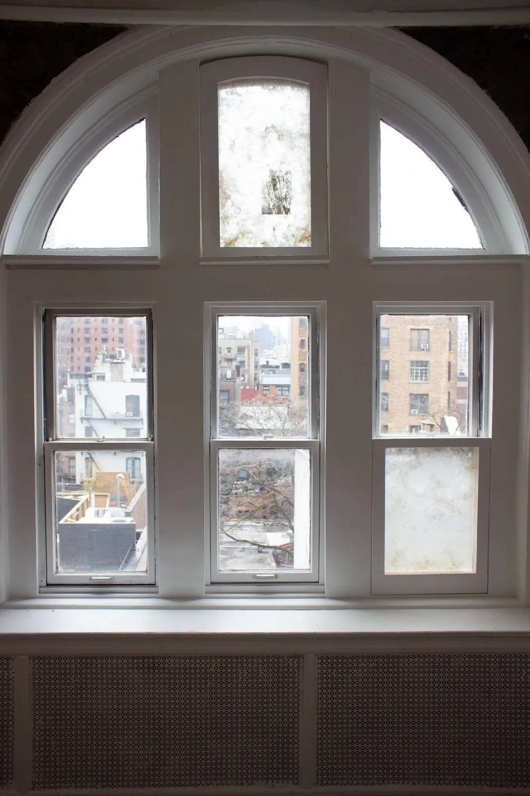An arched window with thick white sills and segmented panes features a view of the city. Certain panes are slightly obscured and clouded with white pigment.