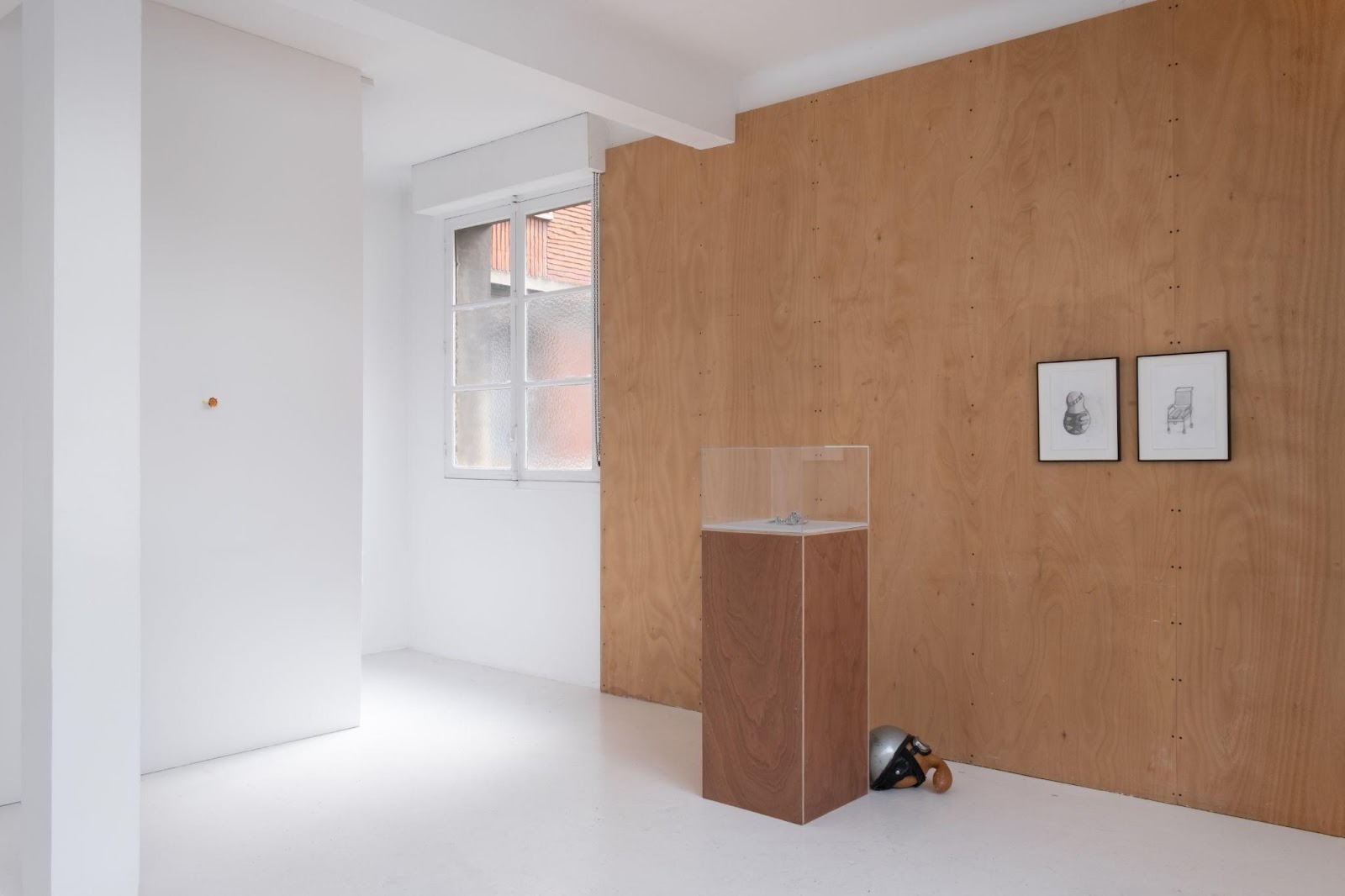Installation view of art exhibition with white walls and wooden walls, two picture frames hang to the right hand side and there's a wooden pedestal with a glass vitrine in the middle.