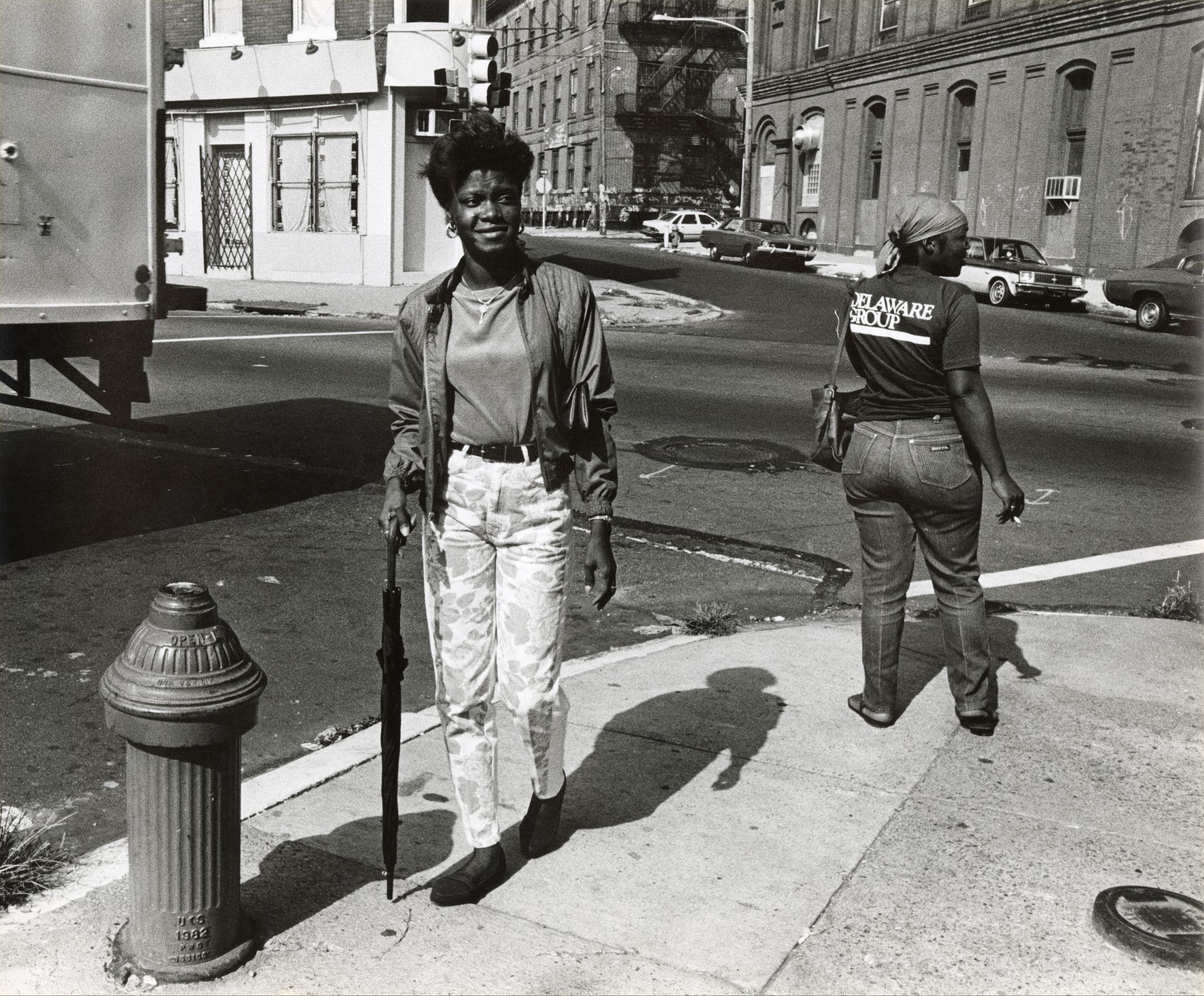 Woman standing on a city sidewalk near a fire hydrant, holding a closed umbrella, smiling at the camera. Another woman walks away in the background. Philadelphia, early 1980s.