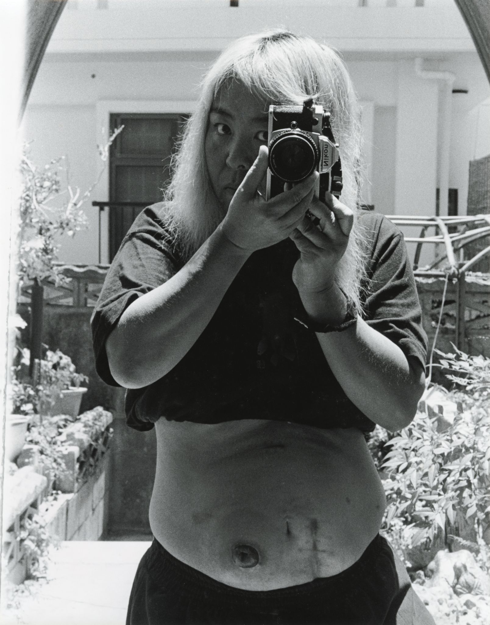 Mirror self-portrait of a woman photographing herself with a Nikon camera, her midriff exposed, surrounded by indoor plants.