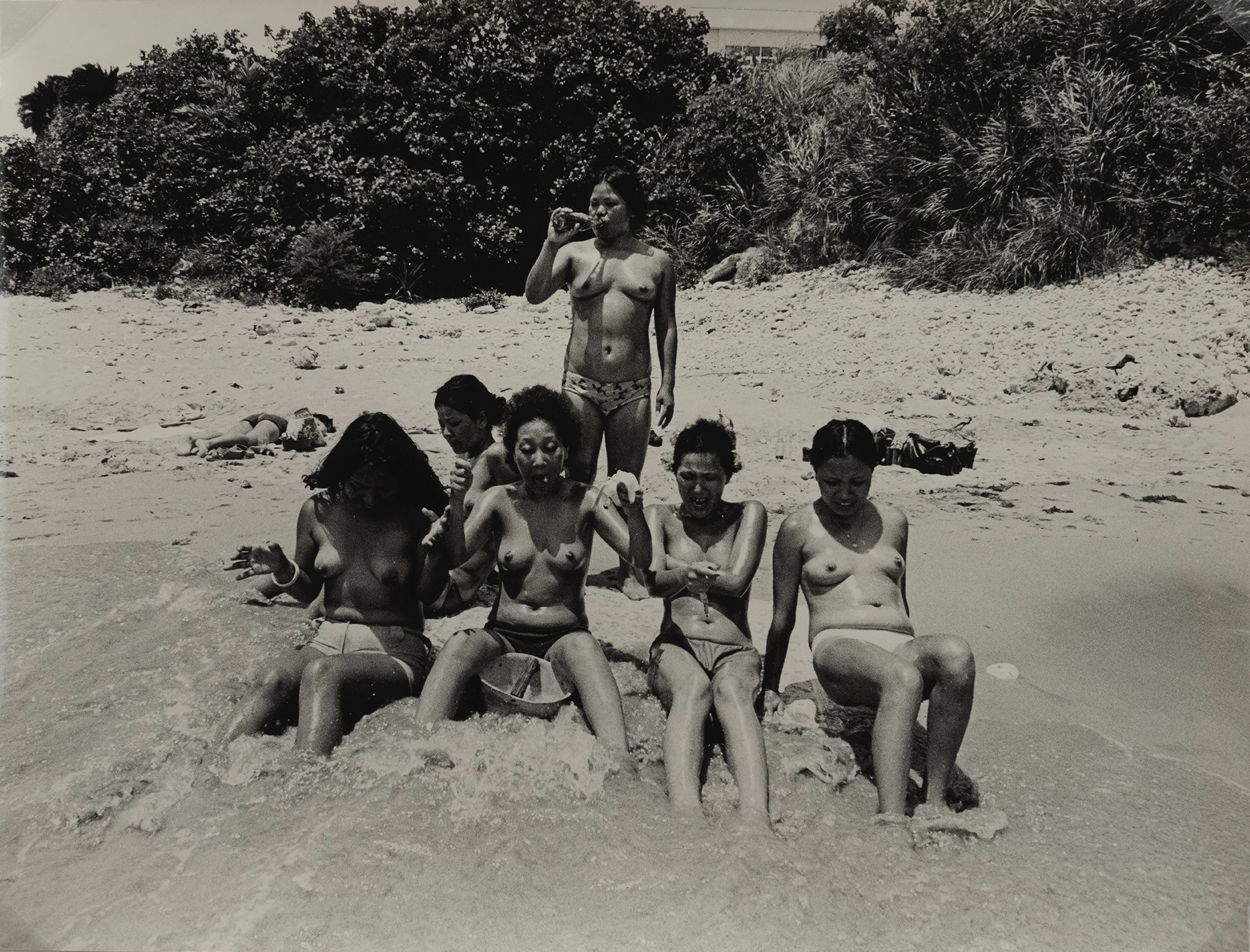 Group of women sitting topless in shallow beach water, one standing behind them drinking from a bottle, lush vegetation in the background.