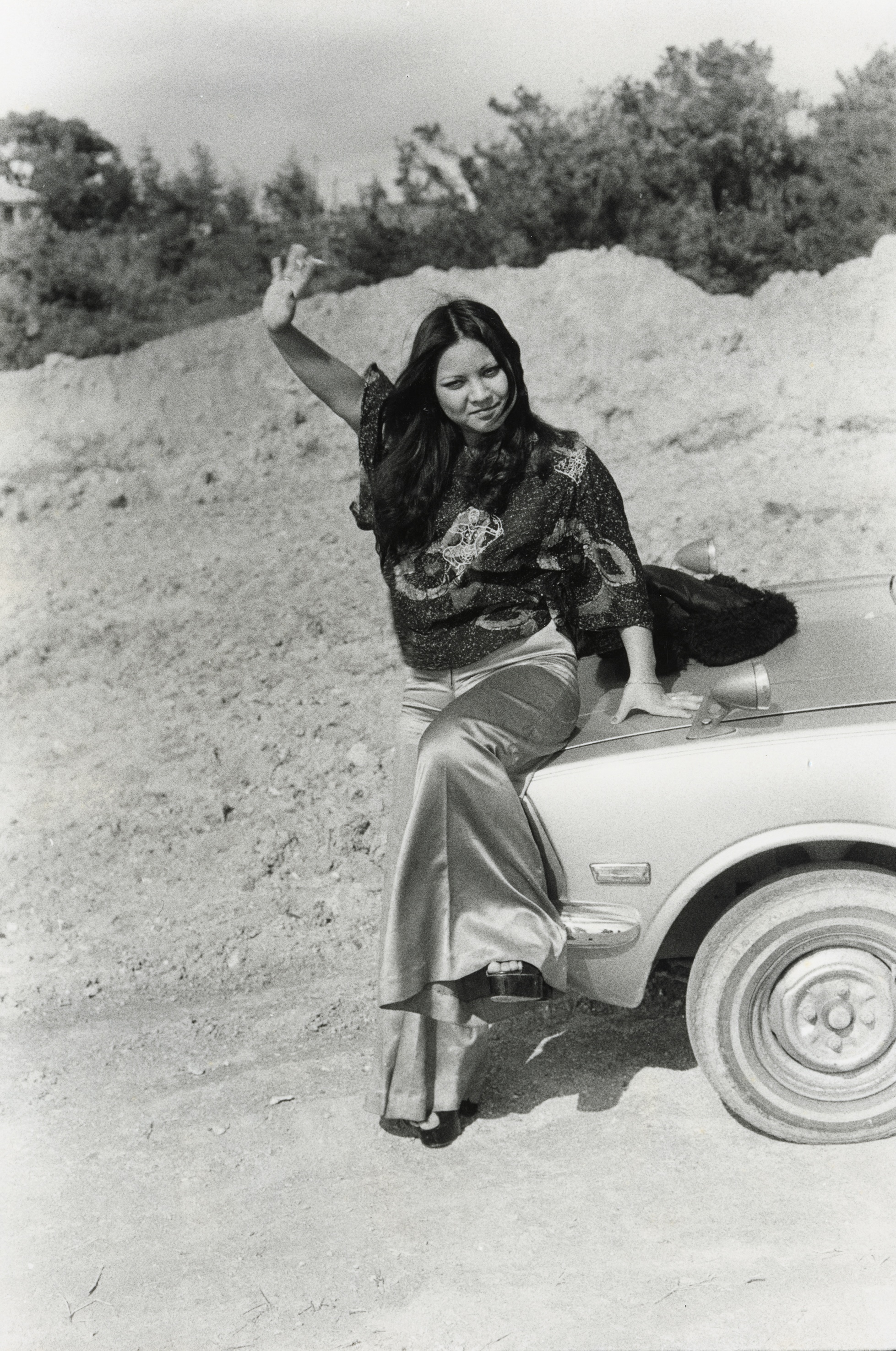 Young woman with long dark hair leaning against the hood of a car in a sandy, open landscape, one arm raised, wearing wide-leg trousers.