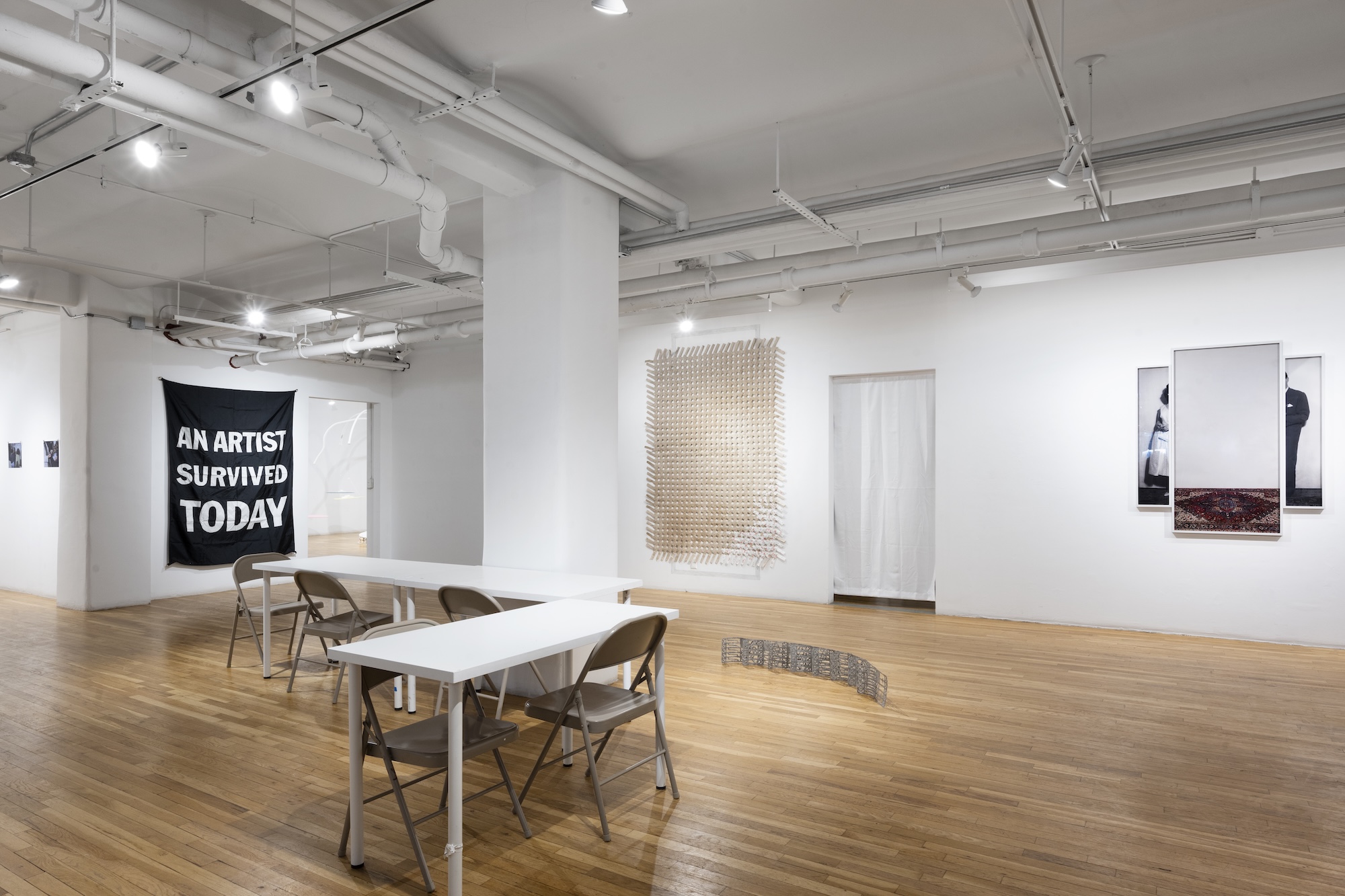 Minimalist gallery with folding tables, chairs, and contemporary artworks, including a black banner reading “AN ARTIST SURVIVED TODAY.”
