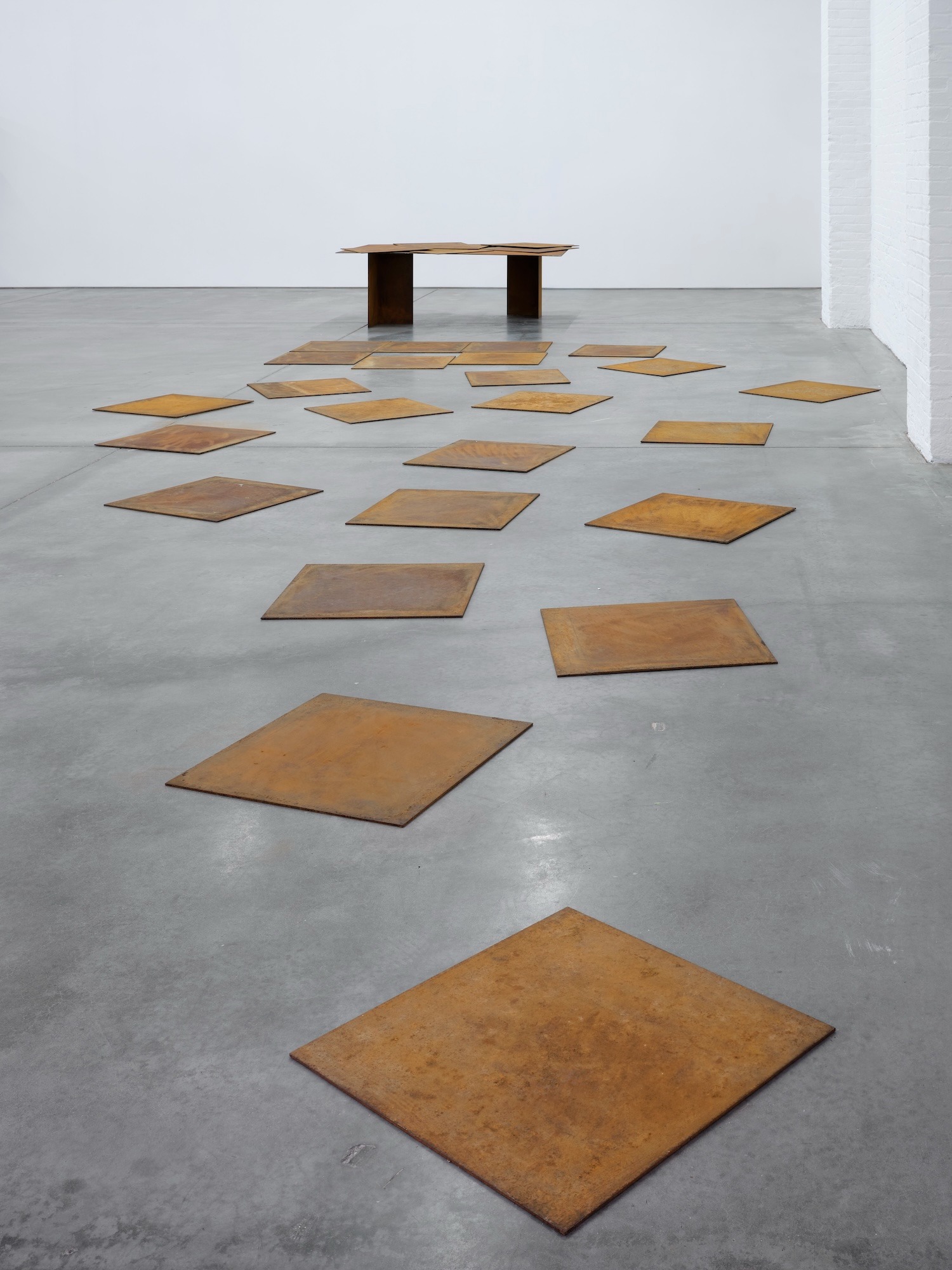 Rusted metal plates scattered across concrete floor around low table sculpture.