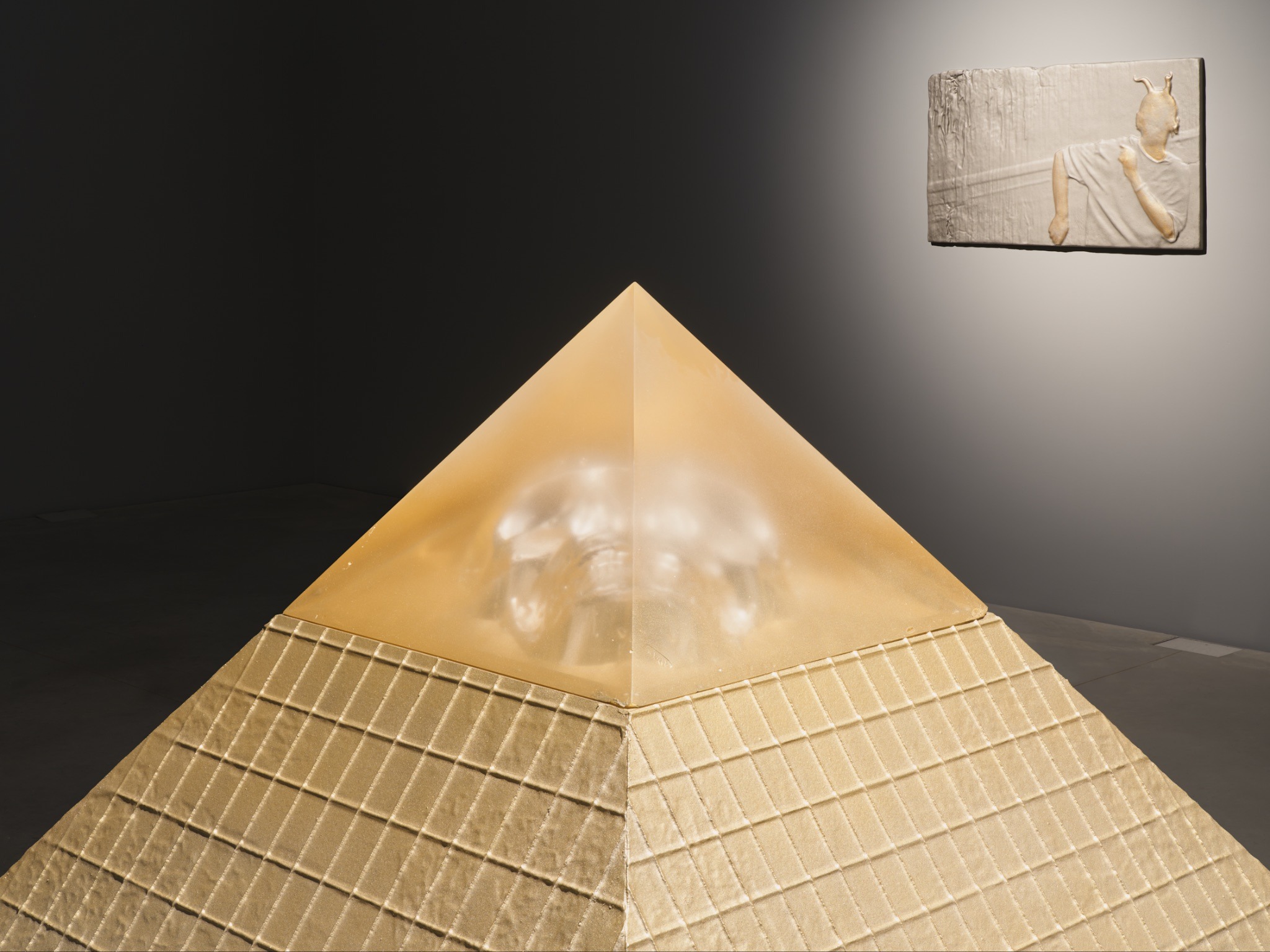 Gold-toned pyramid sculpture with translucent top revealing a face, displayed beside a small relief on a dark gallery wall.