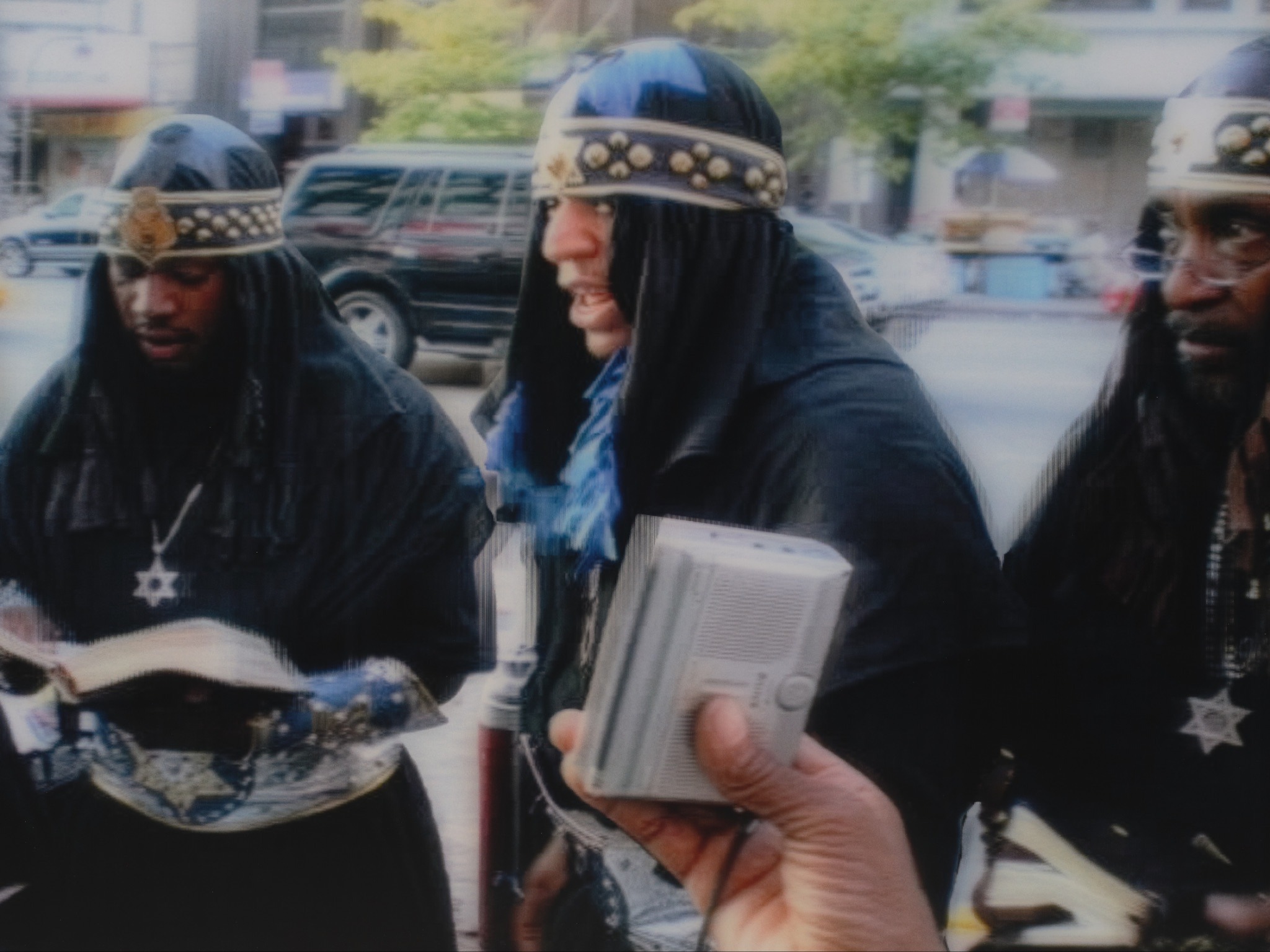 Three men in black robes and studded headbands read from a book on a city sidewalk as a hand holds a small radio.