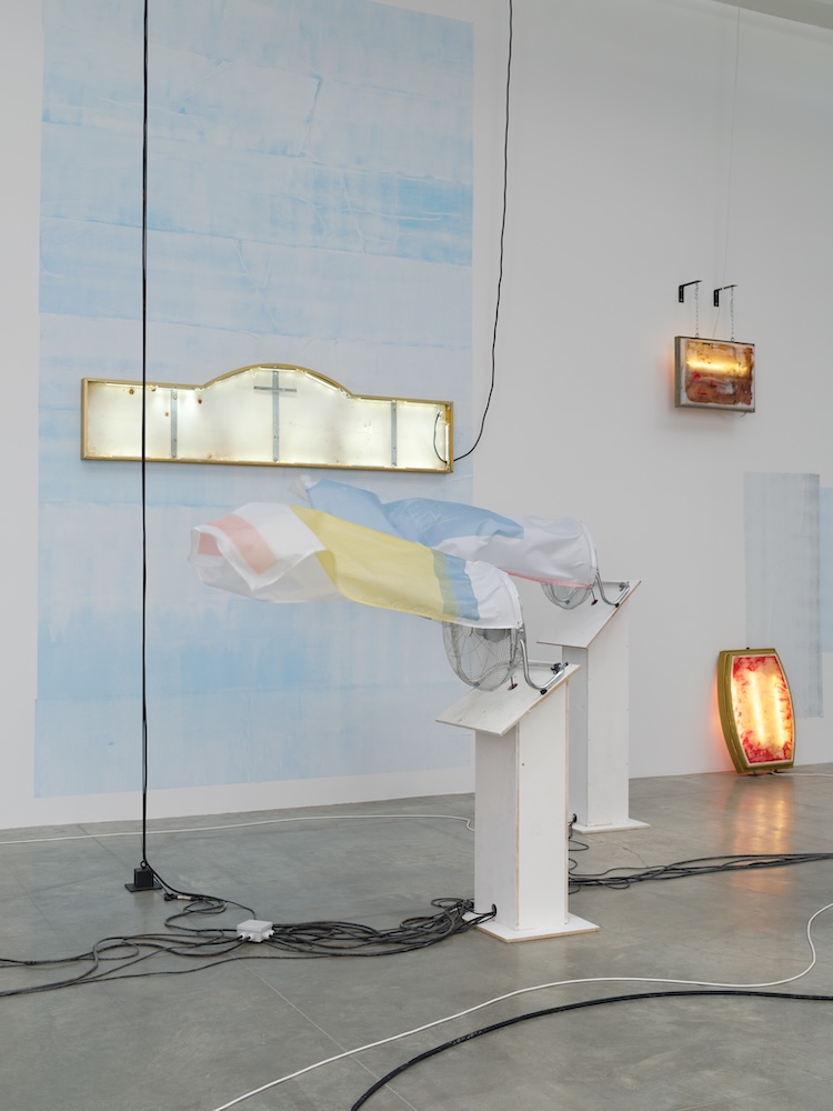 Close installation view of pastel fabric-covered sculpture beside illuminated wall works and pale blue painted panels.